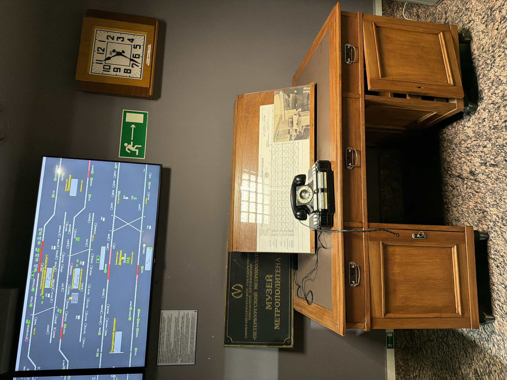 The image depicts a museum exhibit, likely in a historical setting related to transportation. In the foreground, a vintage wooden desk with leather inlay is positioned centrally. On the desk sits a black rotary telephone and a wooden stand displaying a train schedule and a black and white photograph of individuals working in a similar environment. To the left of the desk, a dark plaque with gold lettering reads "САНКТ-ПЕТЕРБУРГСКОЕ ГОСУДАРСТВЕННОЕ УНИТАРНОЕ ПРЕДПРИЯТИЕ «ПЕТЕРБУРГСКИЙ МЕТРОПОЛИТЕН» МУЗЕЙ МЕТРОПОЛИТЕНА," indicating the "Museum of the St. Petersburg Metro."
Behind the desk, a large, wide monitor displays a complex diagram, seemingly a train route map with station names and track layouts in Russian. To the right of the monitor, a square wooden clock with Roman numerals hangs on the wall, displaying the time as approximately 12:54. Below the clock, a green "exit" sign with a running figure is visible. The walls are a neutral grey, and the floor has a granite-like speckled pattern. The overall impression is of a control room or administrative office from a past era, preserved as an exhibit.