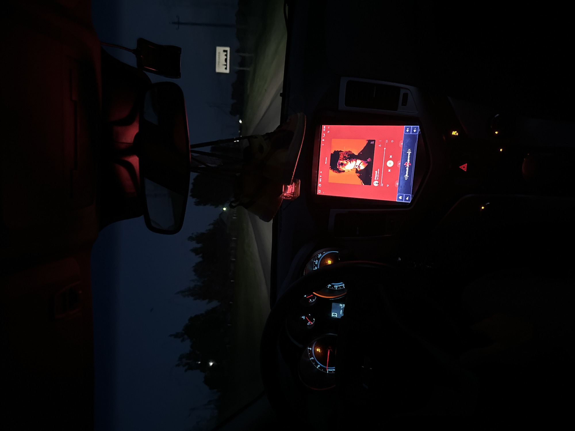 This media file captures the interior of a vehicle at night, likely in a rural setting. The dominant subject is the car's dashboard, illuminated by the glow of its screens and gauges. A large touchscreen display in the center shows what appears to be a music application with album art of a person with glasses and dark curly hair, set against an orange and red background. The track title and artist name are partially visible on the screen, but the text is too blurry to read clearly.
The dashboard features various illuminated gauges and buttons, including a speedometer and tachometer, with some warning lights showing. A hazard light symbol is visible, suggesting the car might be stopped or signaling. Outside the vehicle, the scene is dark, with hints of trees and a dimly lit road stretching into the distance. A small white sign with Arabic-like script is visible on the side of the road, an unusual detail for Belarus, where the provided location context is set. The overall mood is quiet and introspective, illuminated by the artificial light within the car, contrasting with the darkness of the night. The time of day is clearly night, and the weather appears to be calm, given the lack of visible rain or wind effects. The red glow from a light source on the left side of the frame suggests an interior car light or a light from another vehicle.