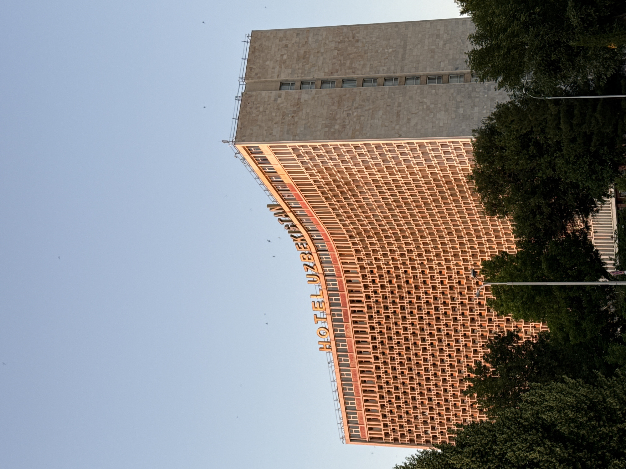 The image depicts the Hotel Uzbekistan in Tashkent, Uzbekistan, viewed from a low angle. The lower portion of the building is covered in a grid-like pattern, with the hotel's name vertically displayed on its facade. Above the patterned section, the building transitions to a plain, textured surface. The scene includes a clear blue sky, suggesting daytime. Trees are visible on the right side of the frame, partially obscuring the lower portion of the building.