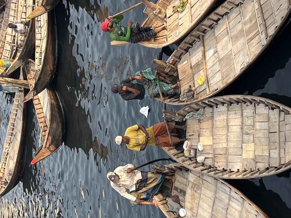 An overhead view captures a busy waterway, likely the Buriganga River in Dhaka, Bangladesh, teeming with numerous traditional wooden boats. The scene focuses on four men in the foreground, each occupying a separate, slender wooden boat with visible floorboards.
From left to right:
1. A man wearing a white plaid shirt, a light-colored cap, and a multi-striped lungi is standing, leaning over the side of his boat, actively pushing a long wooden pole into the dark water. His boat contains two white plastic bowls.
2. Adjacent to him, another man in a yellow t-shirt, white cap, and an orange and brown striped lungi stands in his boat. He holds a small stack of currency in his left hand, looking towards the right. His boat contains various small white and yellow containers.
3. Further right, a man in a dark grey t-shirt, a dark cap, and a green and white plaid lungi is seated on the side of his boat, one leg bent. He rests his right hand on his chin or mouth, appearing to observe. A dark cloth is draped over the boat's edge.
4. The fourth man, in a bright green long-sleeved shirt, a red cap, and a dark plaid lungi, stands in his boat, holding a wooden oar across his left shoulder with both hands. He appears to be looking towards the man in the yellow shirt. His boat contains a yellow plastic bowl and a patterned cloth.
Several other similar wooden boats, some with red-tipped bows, are visible in the background, floating on the dark, rippling water. The boats in the background also contain indistinct figures. The overall impression is of a lively water-based transport or marketplace activity under daylight conditions.