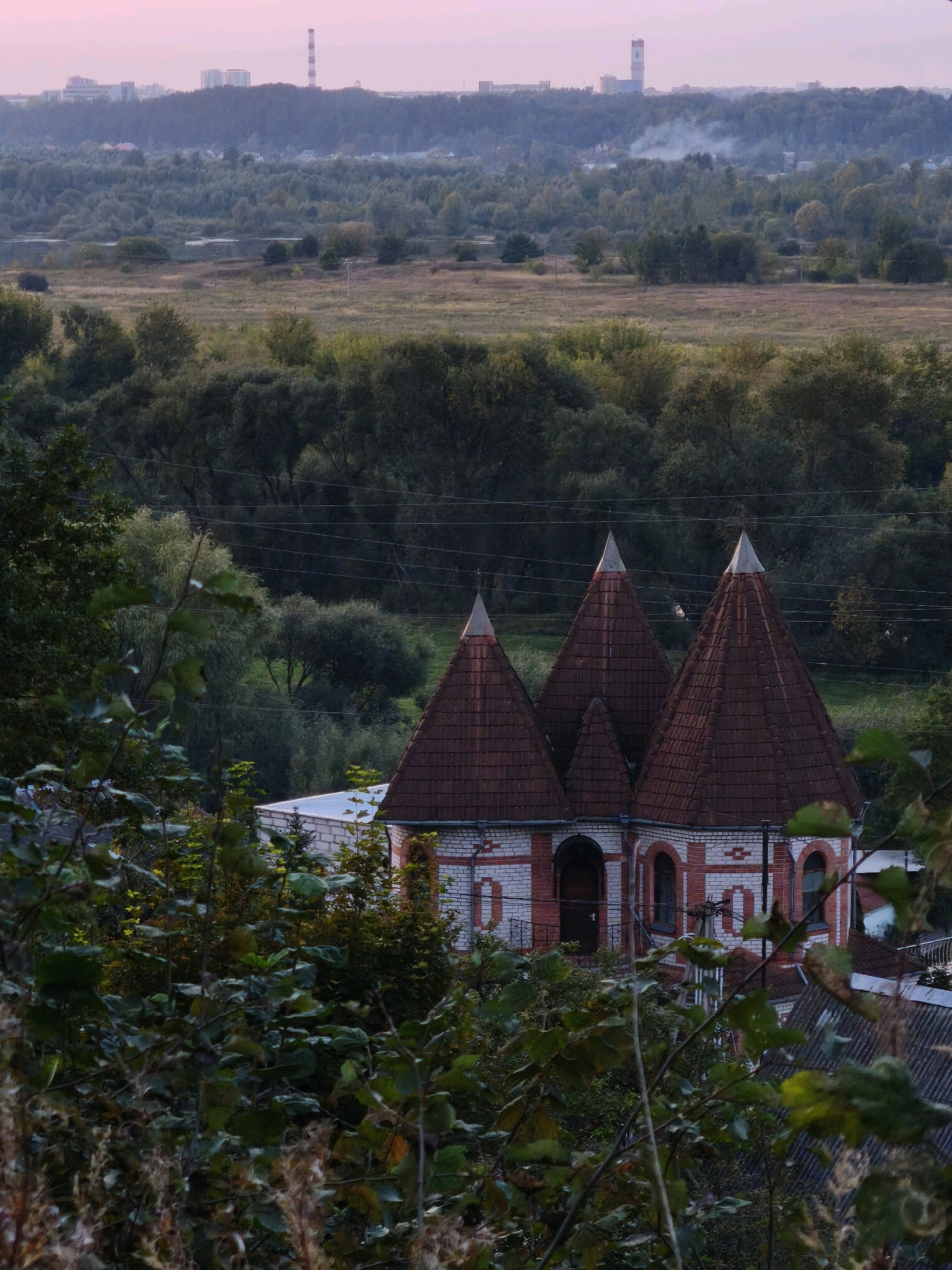 The image depicts a panoramic view of a landscape with a distinctive architectural structure in the foreground, located in the city of Mahilyow, Belarus. The sky is a soft pink, suggesting either dawn or dusk. In the far distance, a city skyline is visible, characterized by several buildings and a tall industrial chimney emitting a plume of smoke.
The mid-ground consists of a rolling landscape covered in dense green foliage and trees, with hints of a river or lake. The terrain appears somewhat uneven, with areas of open fields. Power lines stretch horizontally across the scene, disrupting the natural landscape.
In the foreground, a unique building with three conical, tiled roofs dominates the lower portion of the image. The building's facade is constructed of white and red bricks, with arched windows and doorways. The surrounding area is overgrown with lush green vegetation, with branches and leaves obscuring parts of the structure. The overall impression is one of a rural or semi-rural setting within a city, characterized by a blend of natural elements and human-made structures.