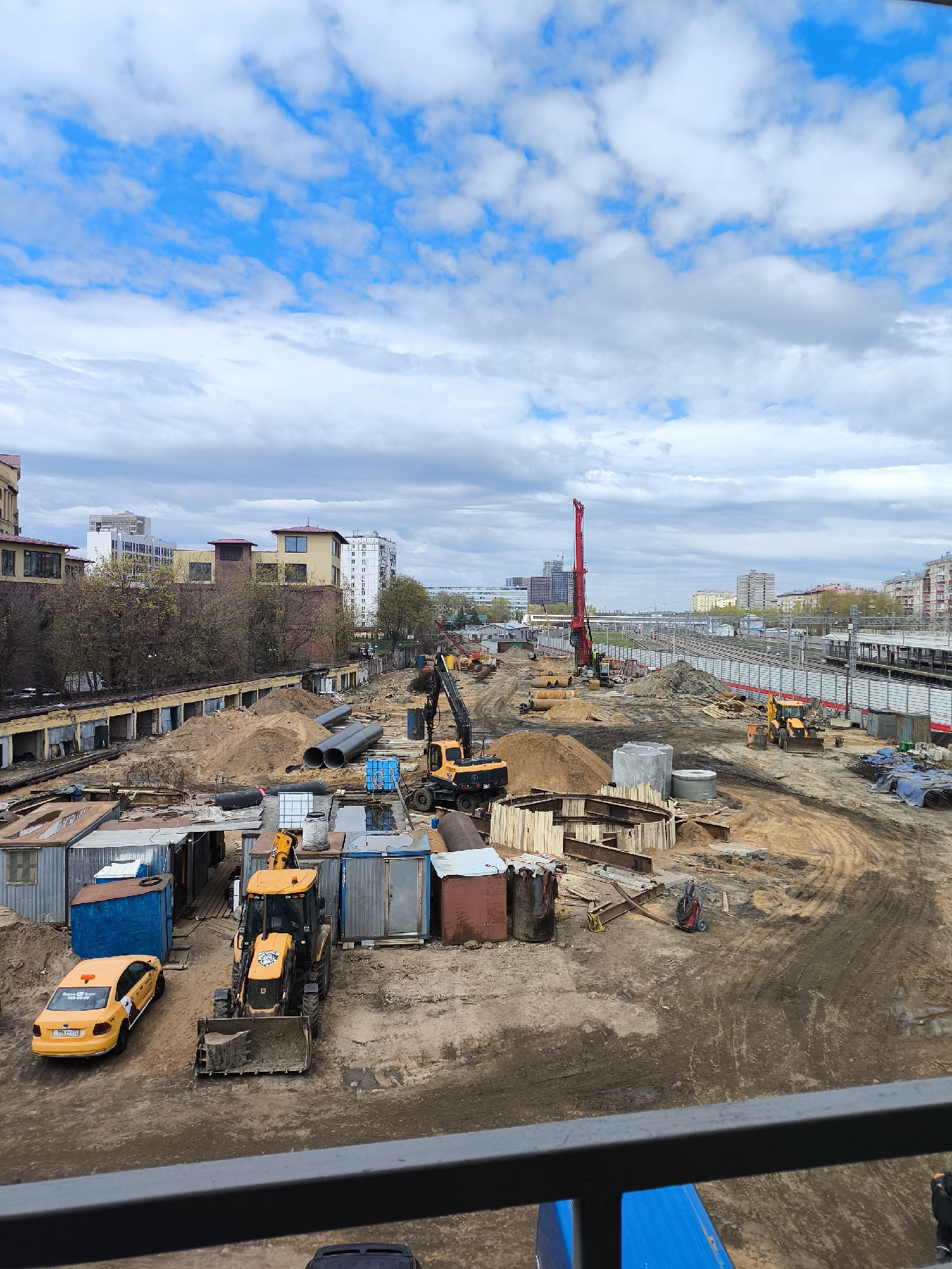 This photo depicts a construction site in Moscow, Russia. Various construction vehicles, including excavators and backhoes, are present on the site, along with materials such as pipes and wooden forms. Temporary structures like storage containers or portable offices are visible. The site is bordered by buildings and infrastructure, suggesting it's in an urban environment. A train station is to the right in the background, and a tall red construction crane pierces the skyline. A yellow taxi is parked next to the building structures on the left. The sky is overcast.