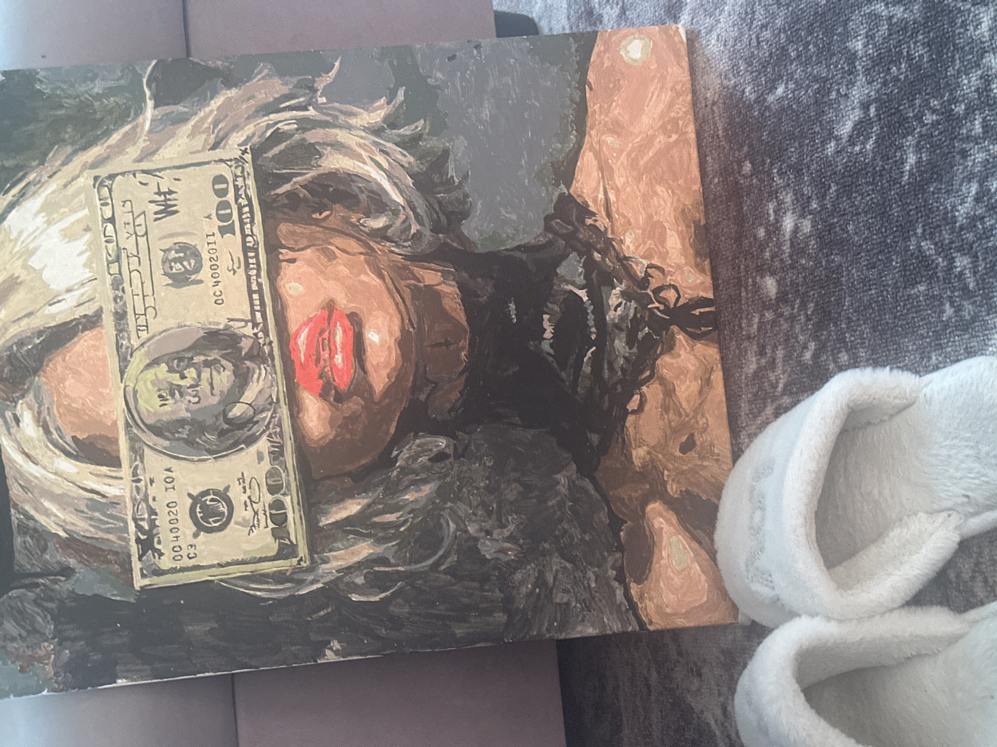 The image shows a painting lying on a grey surface. The painting depicts a person with blonde hair and red lips. A one hundred dollar bill is placed on top of the painting. To the right of the painting is a pair of white fuzzy slippers. There is no indication of location. There is no evidence to support the location being Высокий, Russia.