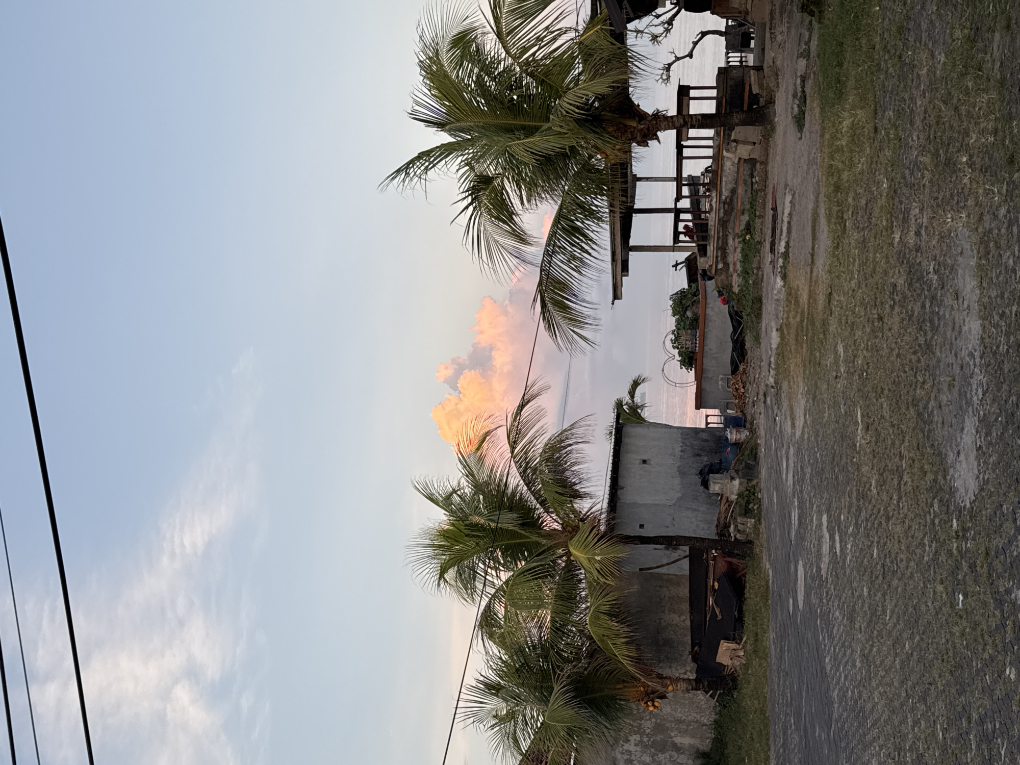 The image shows an outdoor scene in Jimbaran, Indonesia. The sky is a pale blue with scattered clouds tinged with pink. Several palm trees with green fronds are visible. In the background, there is a partially constructed building with wooden support beams. The ground is a mix of pavement, dirt, and grass. Power lines are visible in the upper left.