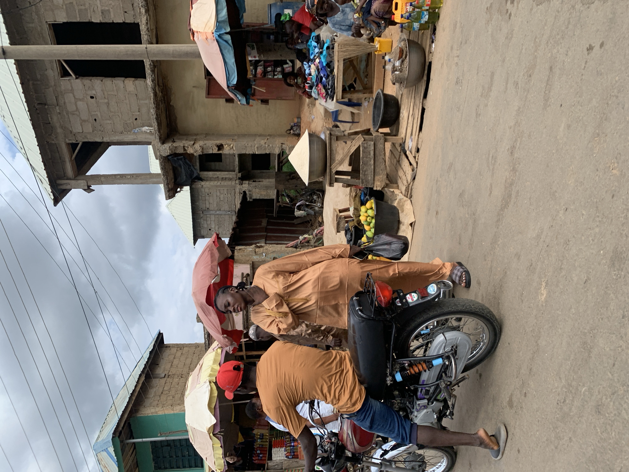 In the city of Akwanga, Nigeria, two men are near a parked motorcycle along a street. One man stands next to the motorcycle, facing the camera, and holding a light red umbrella. He is wearing a light brown robe. The second man is on the motorcycle, wearing a red baseball cap, a light brown t-shirt, blue jeans, and gray shoes. They are near the curb where various vendors are set up selling merchandise such as food, clothing, and other items. Buildings are in the background. The sky is cloudy.