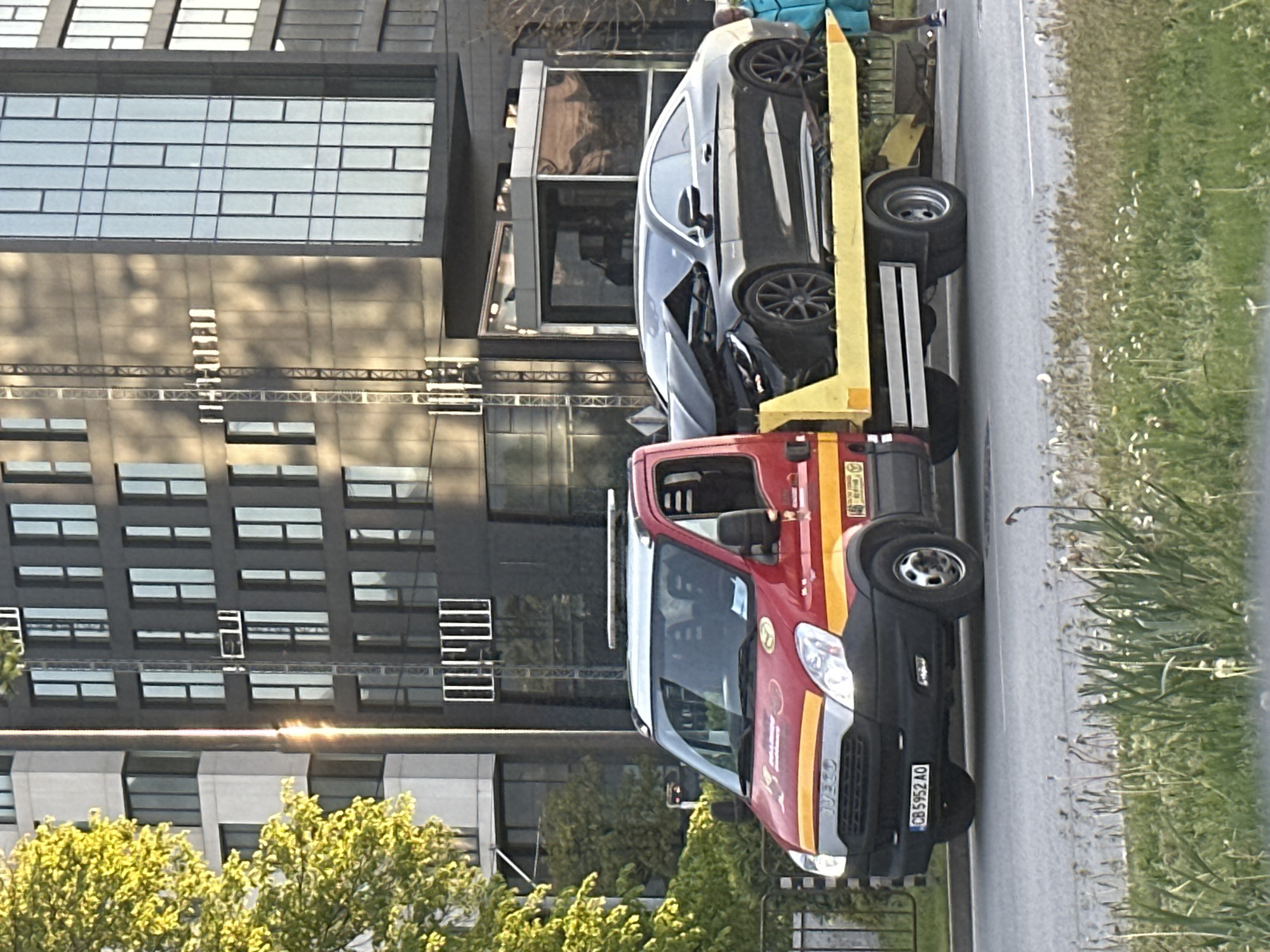 A red and gray tow truck with a yellow flatbed is parked on the side of a road in Sofia, Bulgaria. The truck is carrying a dark gray car. The license plate of the tow truck reads "CB 5952 AO". The background features a modern, multi-story building with many windows. Grass and some trees are visible along the roadside.