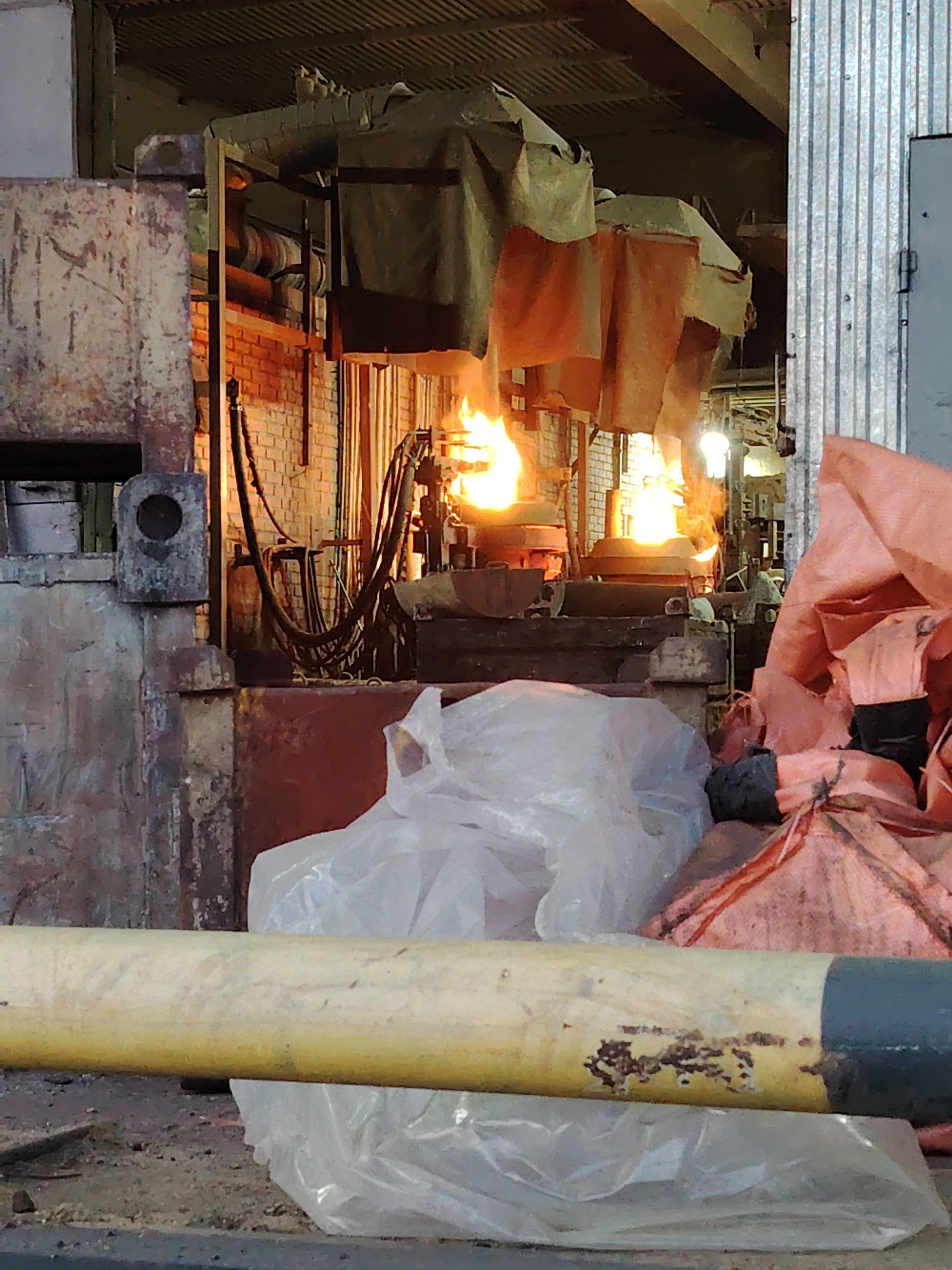 The image depicts an industrial setting, likely a metalworking factory, with molten metal casting and related equipment. Two furnaces with visible flames are present behind protective curtains. Various metal pipes, tubes, and brickwork structures surround the furnaces. In the foreground are a yellow and black painted pipe, discarded plastic sheeting, and other unidentified industrial materials. The location, specified as Noviy, Russia, suggests a potentially outdated or derelict industrial facility. No people are visible in the scene. The setting is cluttered and dimly lit, typical of heavy industrial environments.