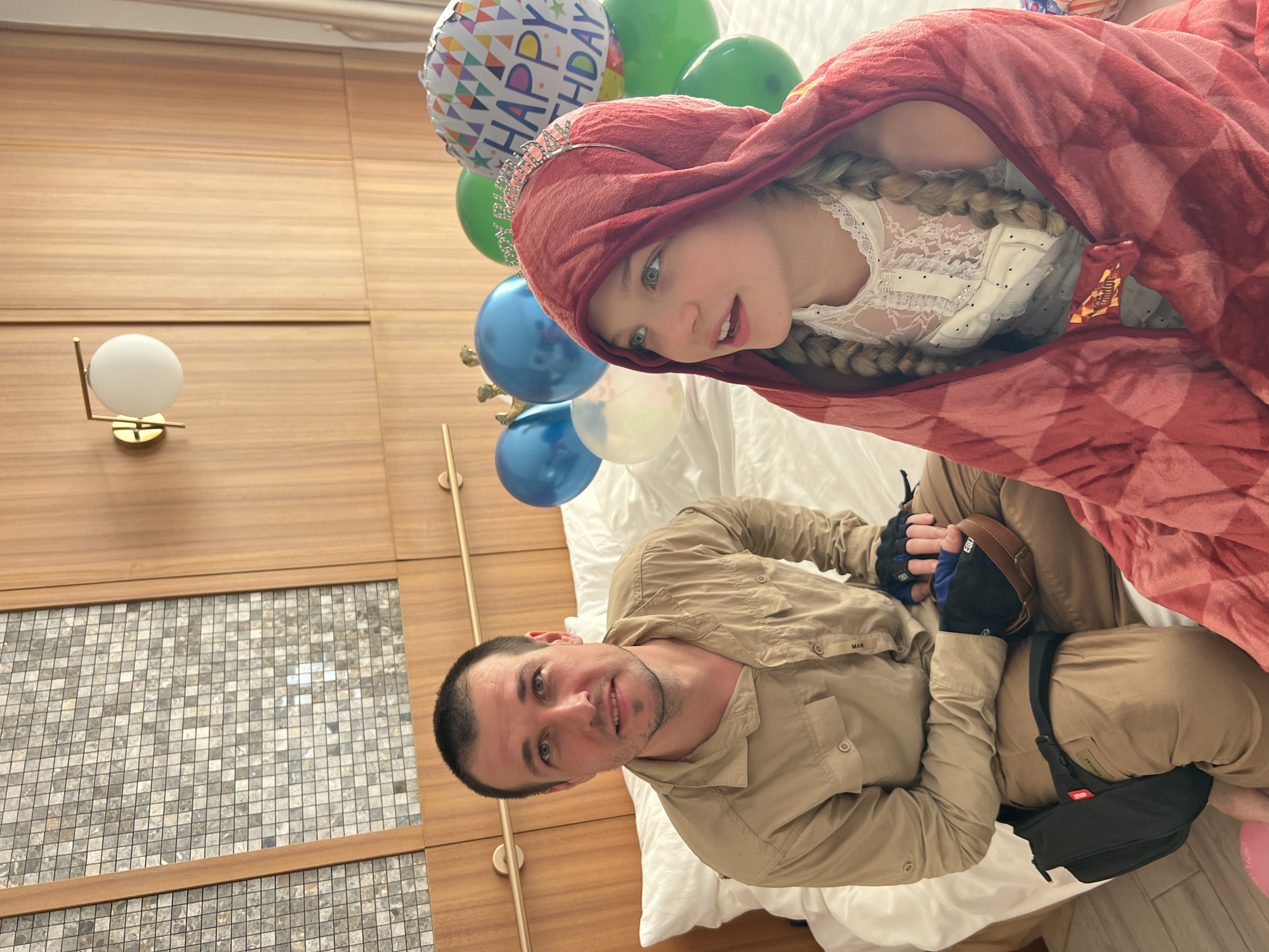 The image shows a man and a young girl, possibly father and daughter, in what appears to be a hotel room or a similar indoor setting. The girl is wearing a red hooded blanket with a crown on her head, suggesting a costume or a celebratory theme. She has long blonde braids and is wearing a white lace top. She is looking towards the camera with a playful expression. The man is wearing a tan button-down shirt and tan pants. He is looking at the camera with a smile.
Several balloons are visible in the background, including a "Happy Birthday" balloon, indicating that a birthday celebration is taking place. The room has wooden paneling on the walls and a bed with white bedding. A modern lamp is mounted on the wall. The overall mood of the image is happy and festive. The lighting suggests it might be daytime, but it's difficult to determine the exact time of day or weather conditions from the image.