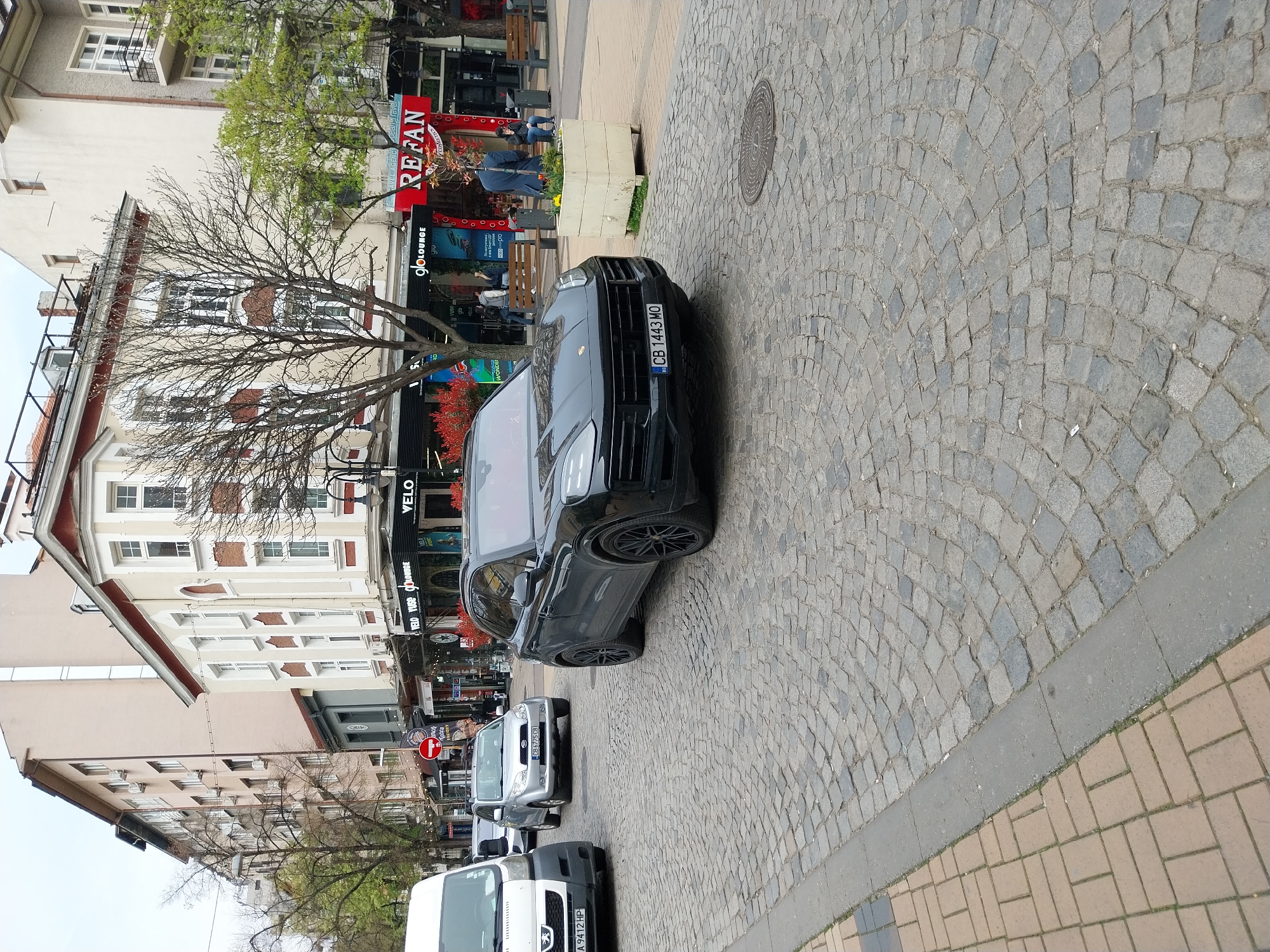 The image depicts a street scene in the city of Sofia, Bulgaria. A black Porsche SUV with the Bulgarian license plate "CB 1443 MO" is parked on a cobblestone street. Behind the Porsche, there are several other parked vehicles including a gray Suzuki and a white van with the license plate "A 9412 HP". Buildings line the left side of the street, with shops and businesses visible. The storefronts include "REFAN COSMETICS", "gloLOUNGE", and "VELO". A leafless tree stands near the buildings. People are visible in the background near the storefronts. The pavement transitions from cobblestone to a sidewalk made of rectangular pavers. A manhole cover is visible on the cobblestone street. A "No Entry" traffic sign is visible near the shops.