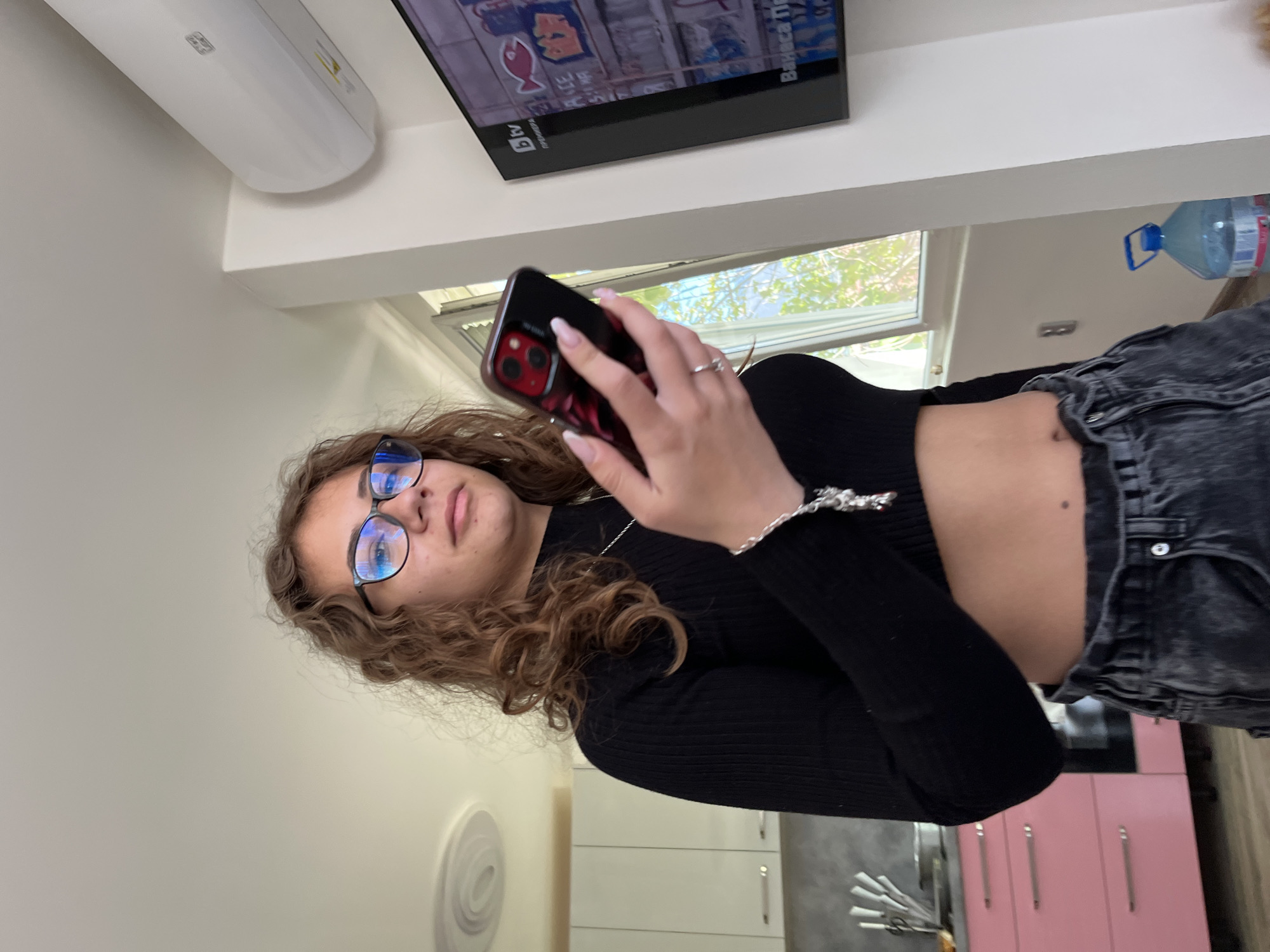A young woman with curly hair and glasses holds a smartphone in a kitchen. She is wearing a black long-sleeved top and grey jeans. The kitchen has white and pink cabinets and is illuminated by natural light coming through a window. There's an air conditioning unit and a television on the wall. A water bottle is on the upper right portion of the image. The city of Karavelovo, Bulgaria, cannot be verified from this image.