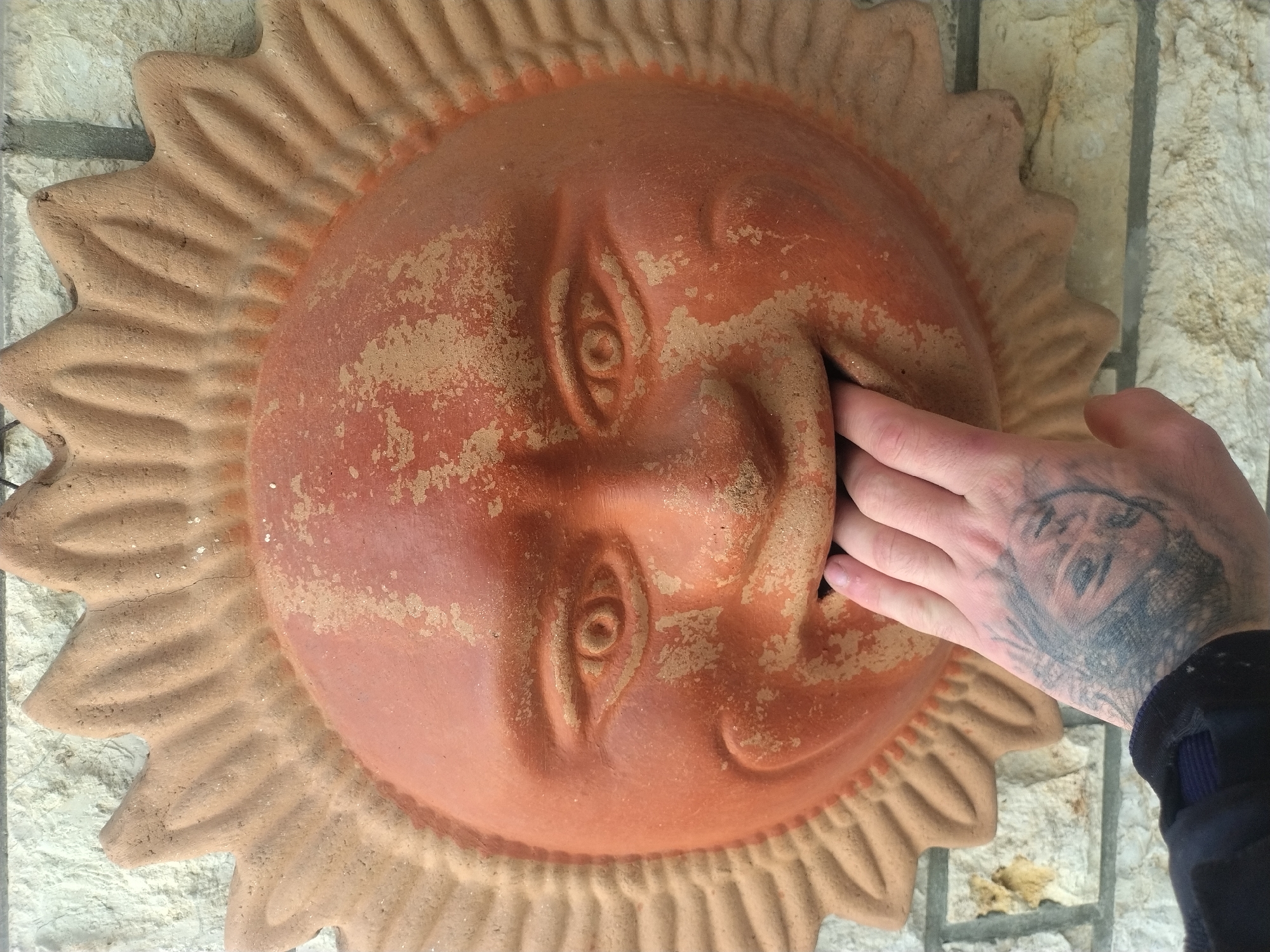 A medium shot depicts a light-skinned hand with a tattoo on the back, fingers inserted into the mouth of a terracotta sun face mounted on a stone wall. The terracotta face has a circular design with stylized rays extending outward, and the facial features are expressive with eyes, nose, and a slightly open mouth. The terracotta has a reddish-brown color with variations in tone and texture, suggesting weathering or age. The hand appears to be pushing something into the face's mouth. The wall behind the face is constructed of light-colored stone bricks. The location is likely Luxembourg, Luxembourg.