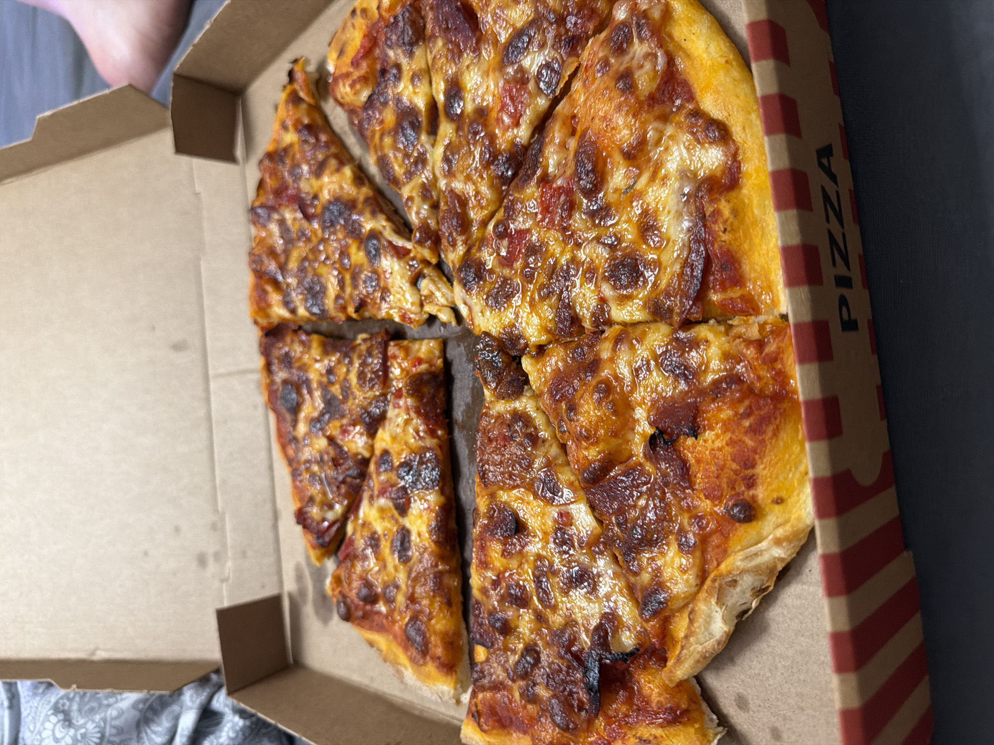 A pizza box containing six slices of pizza is on a dark surface. The pizza is topped with melted cheese and pepperoni. The pizza box has a red and white striped pattern on the inside of the lid and "PIZZA" printed on the outside. The scene is indoors and the lighting is dim. There is no indication of the location in Ruse, Bulgaria.