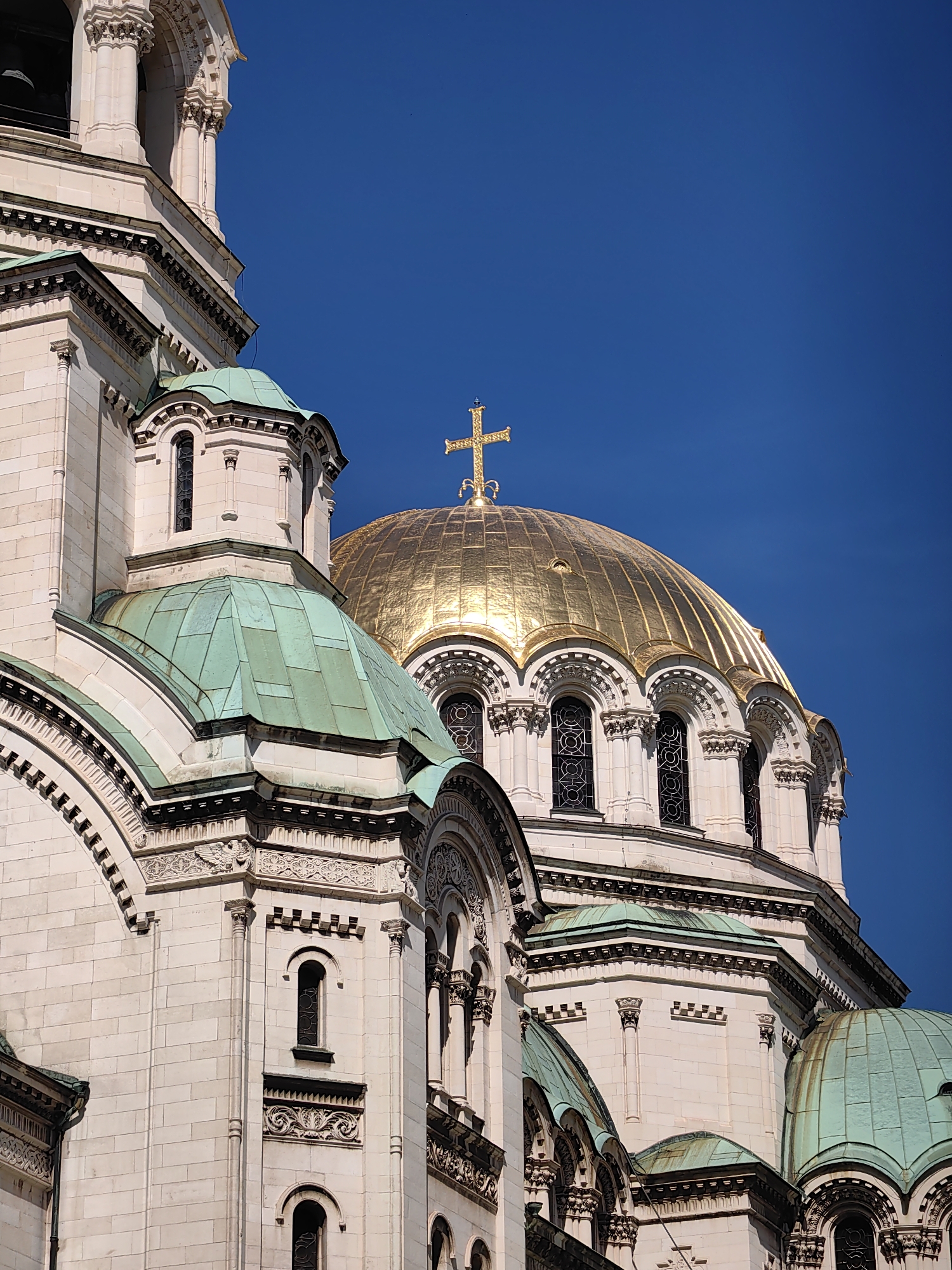This is a close-up, angled exterior shot of the Alexander Nevsky Cathedral in Sofia, Bulgaria, on a clear, sunny day with a vibrant blue sky. The image focuses on the cathedral's striking architecture, featuring cream-colored stone blocks and multiple domes. Prominently displayed is a large, gilded dome topped with a golden cross, reflecting the sunlight. Adjacent to it, a smaller, green-patinaed dome is visible, also with decorative architectural details. The building exhibits intricate carvings, arched windows with decorative grilles, and tiered sections with columns and moldings. The perspective emphasizes the scale and grandeur of the religious structure, with no people or other objects in the frame besides the cathedral and the sky.