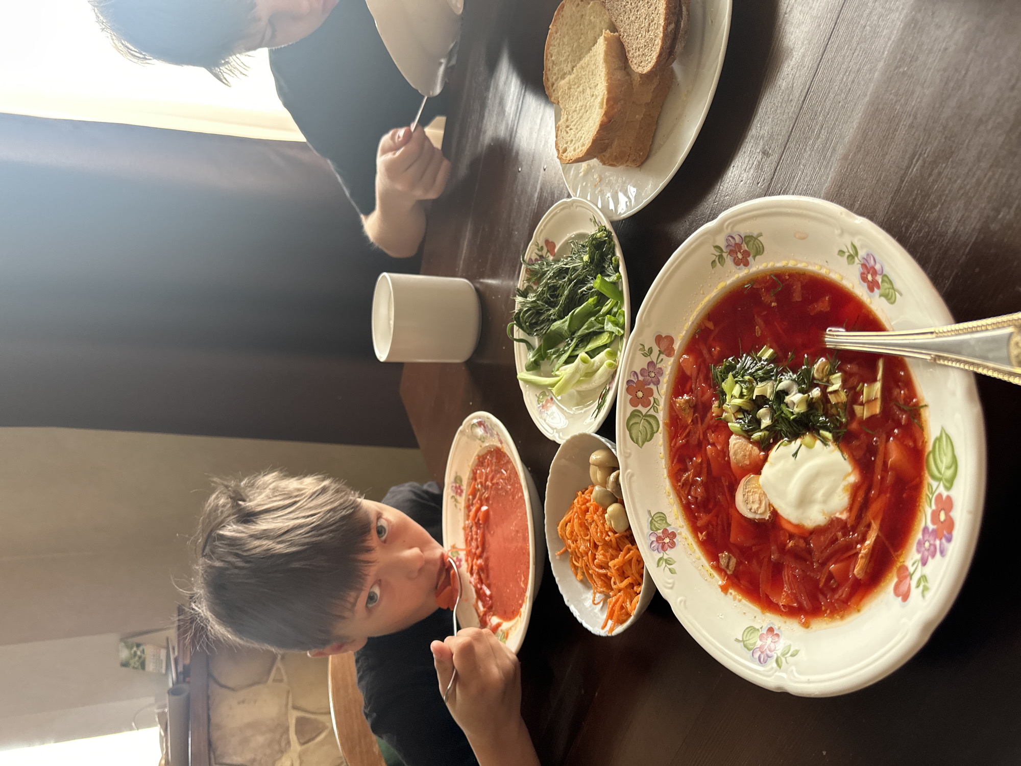 A family is seated at a table, likely in a home in Chaikovsky, Russia, enjoying a meal that includes borscht, rye bread, and various side dishes. One child is in the foreground, eating from a bowl, while an adult is partially visible at the top of the frame. The table is set with floral-patterned bowls and plates, and a white mug is also present.