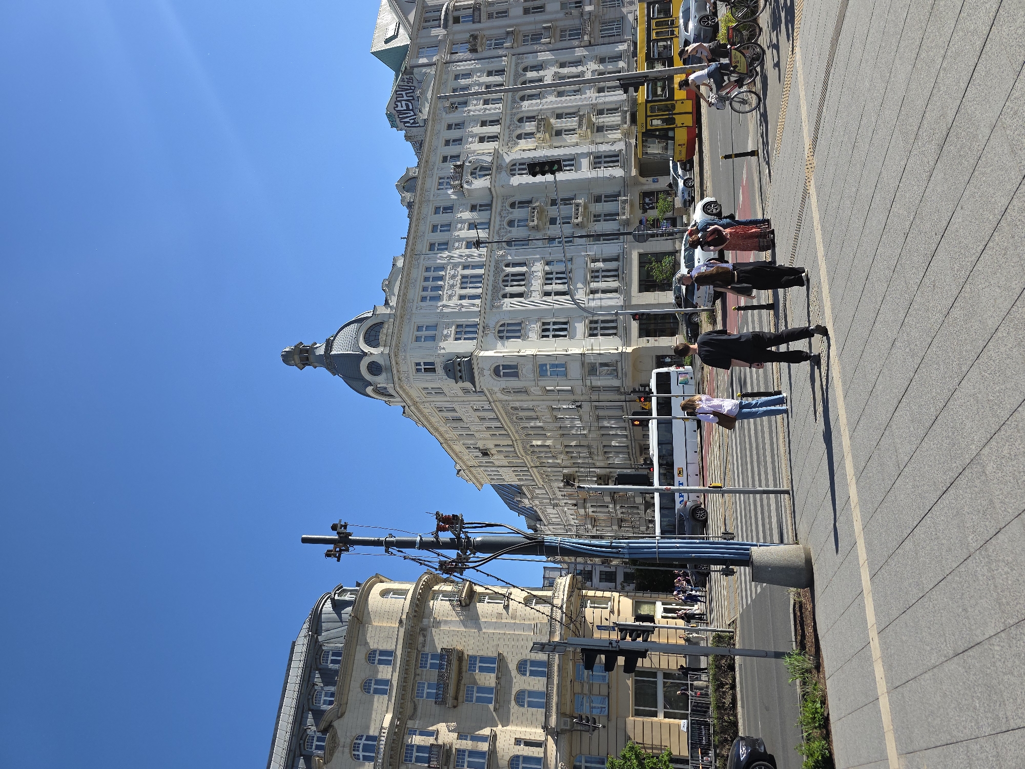 The image shows a street scene in Warsaw, Poland, on a sunny day. A tall, ornate, white building with a dome top is on the left side of the frame. A yellow and red tram is visible behind a group of people standing on the sidewalk. A person on a bicycle rides along the sidewalk next to the tram. The foreground features a paved sidewalk with a concrete curb, and several people are walking and waiting to cross the street. A street lamp pole with wires and traffic signals extends across the view, partially obscuring another building. A parked car and some greenery are at the bottom of the frame. The clear blue sky is visible above the buildings.