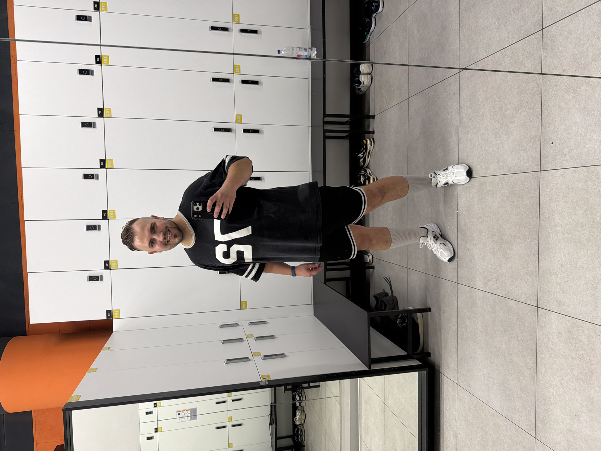 An adult male stands in a well-lit locker room, presumably located within a fitness center in Moscow, Russia. The man, with light skin, short brown hair, and a short beard, is centered in the frame, smiling directly at the camera of a black smartphone (likely an iPhone) held in his right hand, indicating he is taking a selfie. He is dressed in a black oversized short-sleeved t-shirt with large white "SO" lettering on the front, black athletic shorts with white trim, white ankle socks, and white athletic shoes with black accents. A dark-colored smartwatch is visible on his left wrist.
The locker room features white, vertically arranged lockers with black locking mechanisms, marked with yellow numbered stickers such as "191," "192," "193," "194," "195," "196," and "197" on the right side. Black benches are positioned along the base of the lockers. The floor is tiled with light grey squares and dark grout lines. A large mirror covers the entire left wall, reflecting the locker room, including additional lockers, benches, and various pairs of shoes placed underneath. A curved orange pillar and orange ceiling section are visible in the upper left, contrasting with an upper black ceiling area. A clear plastic water bottle with a blue cap rests on a bench to the right of the man.