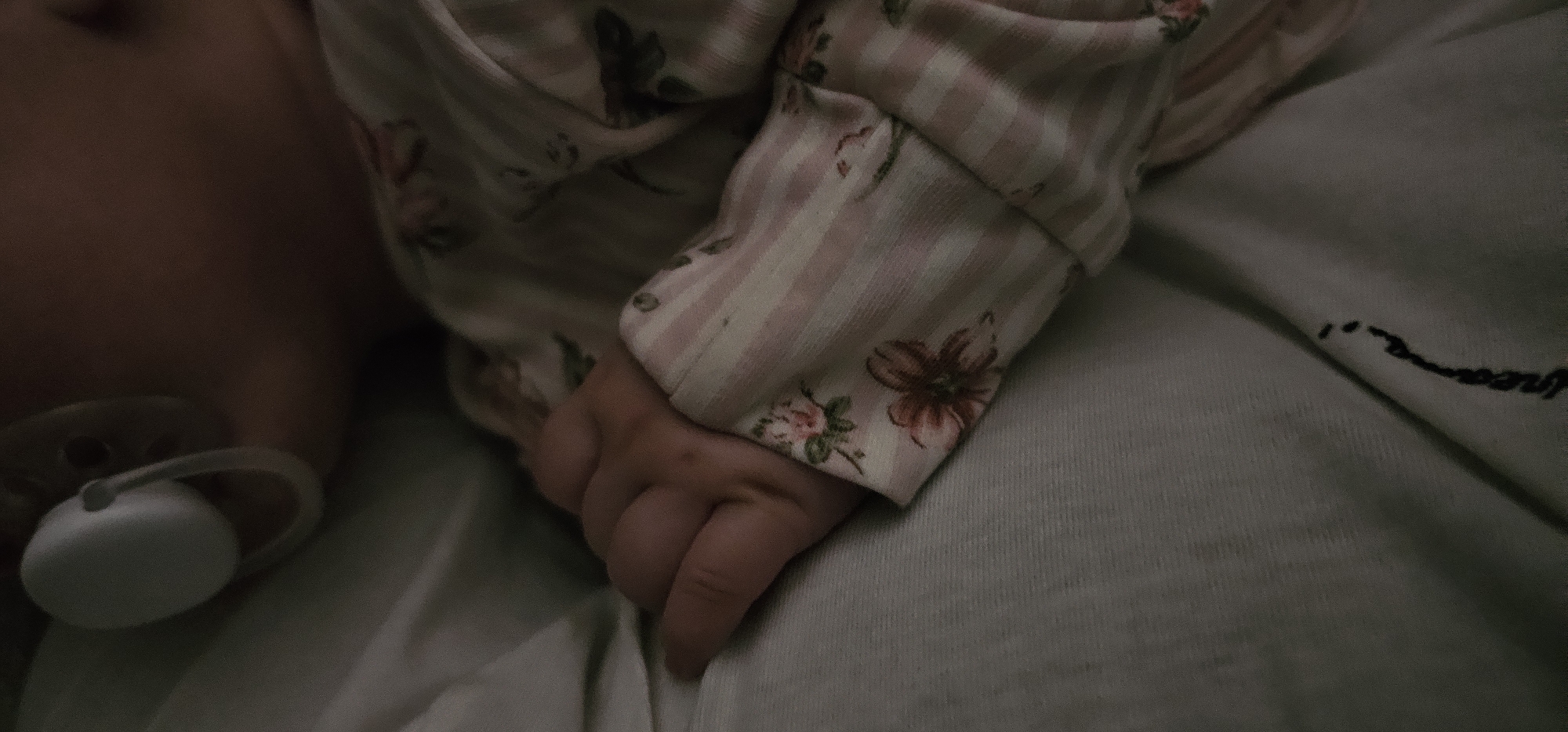 A baby rests on a light-colored surface, possibly a blanket or bed, with a pacifier near their face. The baby is wearing a light pink and white striped onesie with floral accents. The scene is dimly lit.