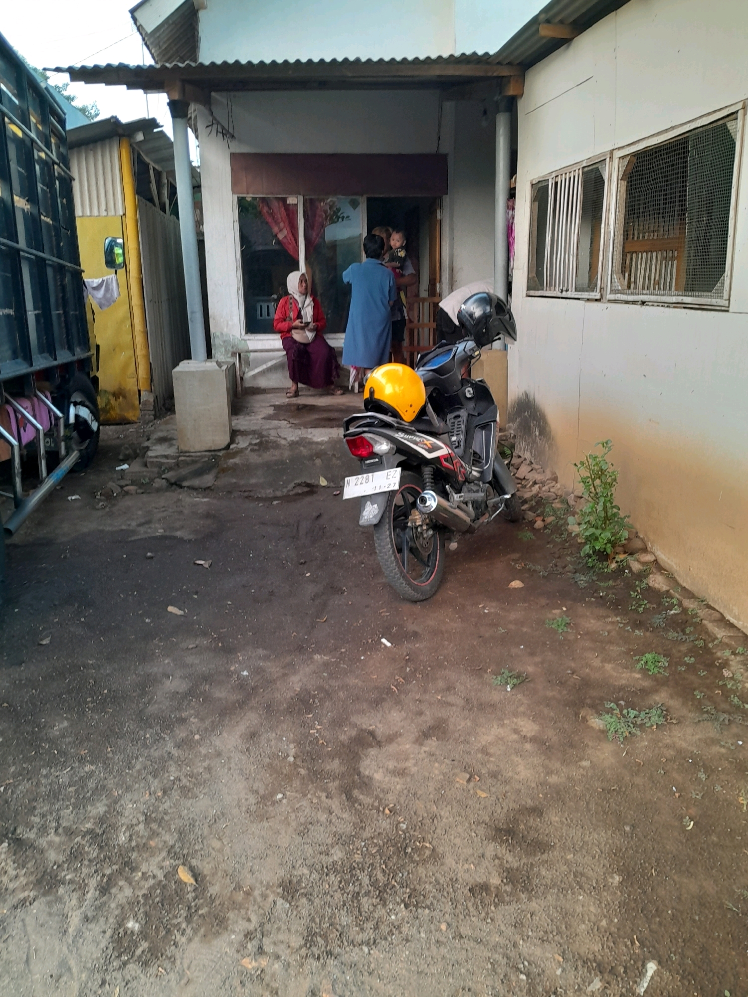 The image depicts an outdoor scene in front of a building with a corrugated metal roof. A black motorcycle with a yellow helmet on its handlebars is parked on a dirt and gravel ground. The motorcycle has a license plate that reads "N 2281 EZ". To the left of the motorcycle, a large truck with a black cargo bed is partially visible. In the background, a woman in a red hijab and dark clothing is seated on a step, holding a mobile phone. Another person in a blue outfit is standing near her. A child is being held by someone further inside the doorway of the building. The building itself has white walls and windows with metal grates. The ground is uneven and appears to be a mix of dirt, small stones, and some sparse vegetation. The overall impression is a casual, everyday scene in what appears to be a residential or small commercial area.