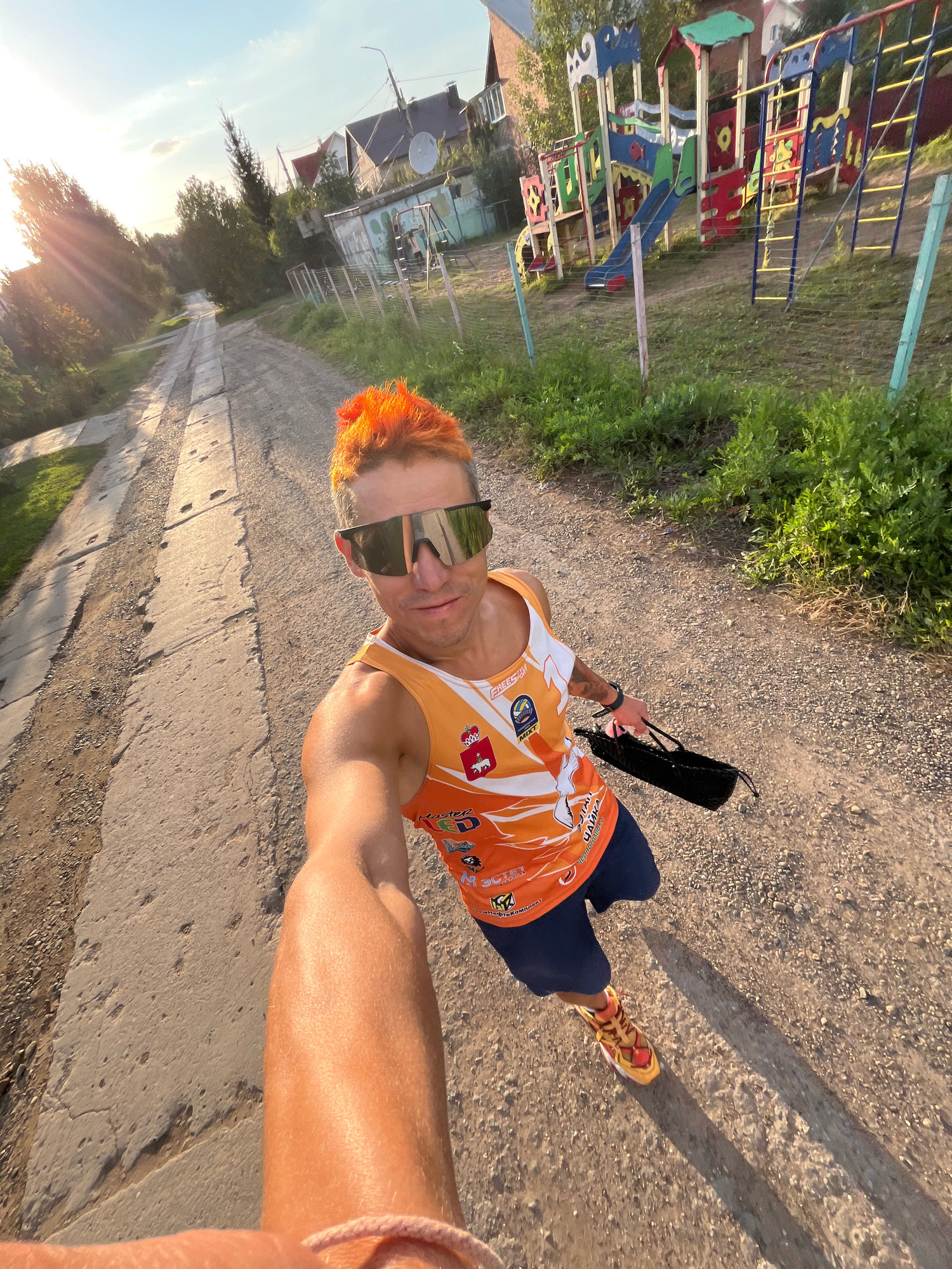 A man with bright orange hair and large sunglasses is taking a selfie outdoors on a sunny day. He is wearing an orange athletic tank top with various logos, dark blue shorts, and colorful running shoes. He is holding a black bag in his right hand. The man appears to be on a dirt path or road with some concrete sections. In the background, there is a playground with colorful structures, houses, and trees. The scene is consistent with a rural or suburban area. The image appears to have been taken in the city of Chaikovsky, Russia, as indicated by logos on the man's shirt.
