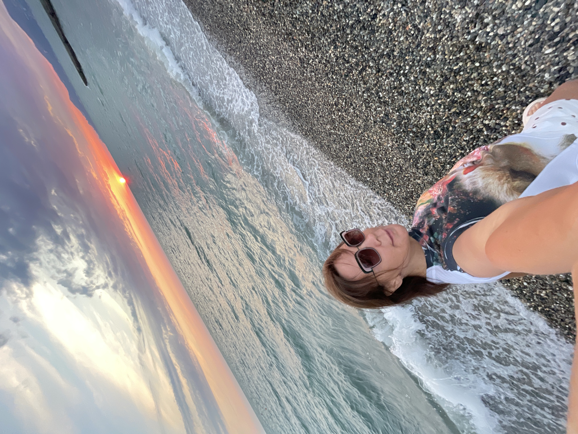 A woman wearing sunglasses and a colorful top takes a selfie on a pebble beach at sunset. The ocean waves are gently lapping the shore. The sky is filled with dramatic clouds in shades of orange, pink, and blue, and the sun is a bright orange orb on the horizon. Mountains are visible in the distance.