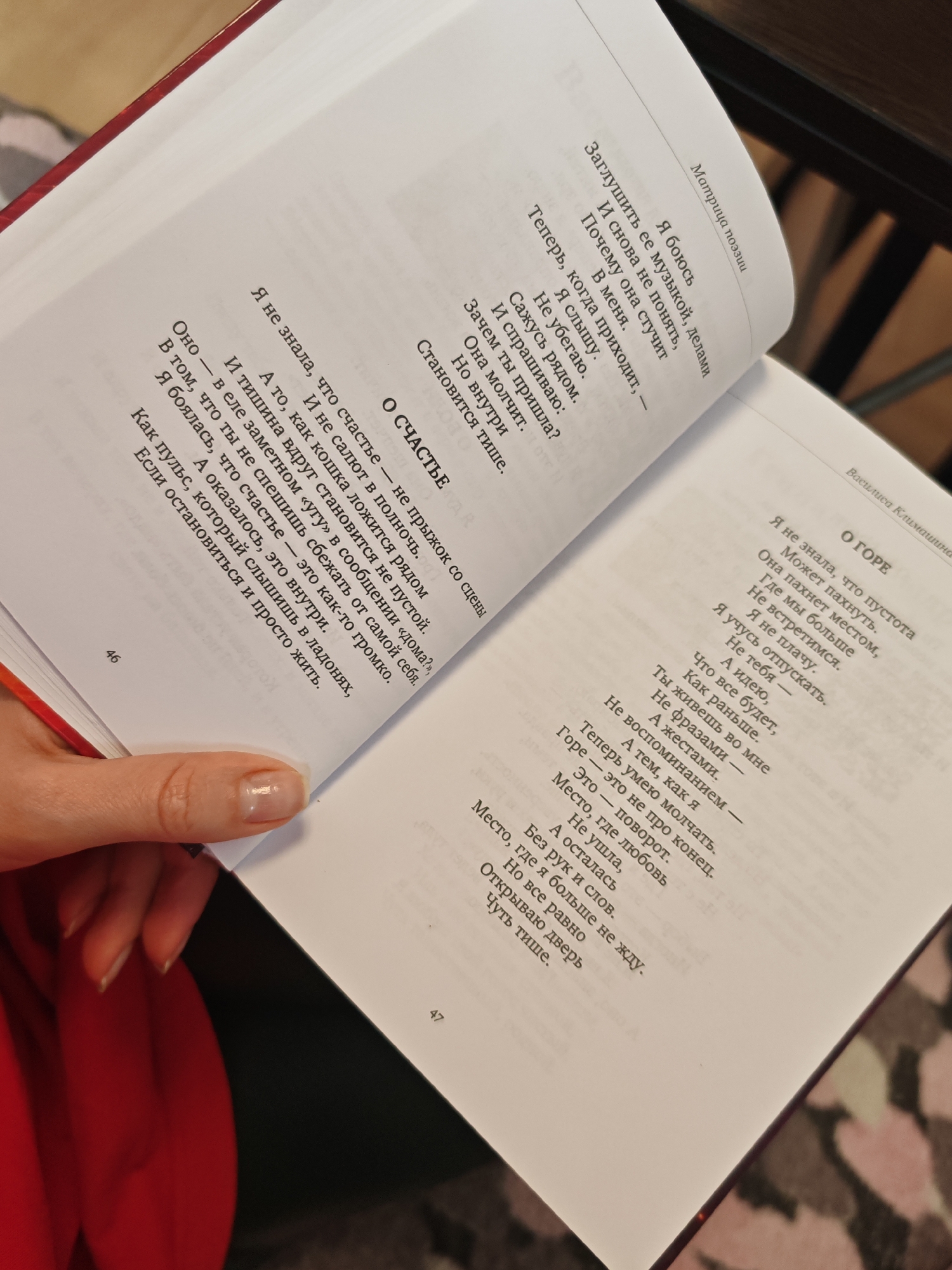 The image shows a close-up of an open book, viewed from above, with a person's hand holding the pages. The book is a collection of poems, with titles like "Матрица поэзии" (Poetry Matrix) and "О СЧАСТЬЕ" (About Happiness) and "О ГОРЕ" (About Grief) visible. The text is in Russian. The background is dark, likely a table or floor, with a hint of a patterned surface visible in the lower right corner. The person's hand is light-skinned with neatly manicured nails, and they are wearing a red garment, possibly a shirt or scarf, which is also visible in the lower left corner. The book is positioned on the dark surface, and the pages are slightly curved. The lighting appears to be from an overhead source, creating shadows on the pages. The image is taken at an angle, with the book's spine and pages creating a diagonal line across the frame. There is no indication of a specific city or location.