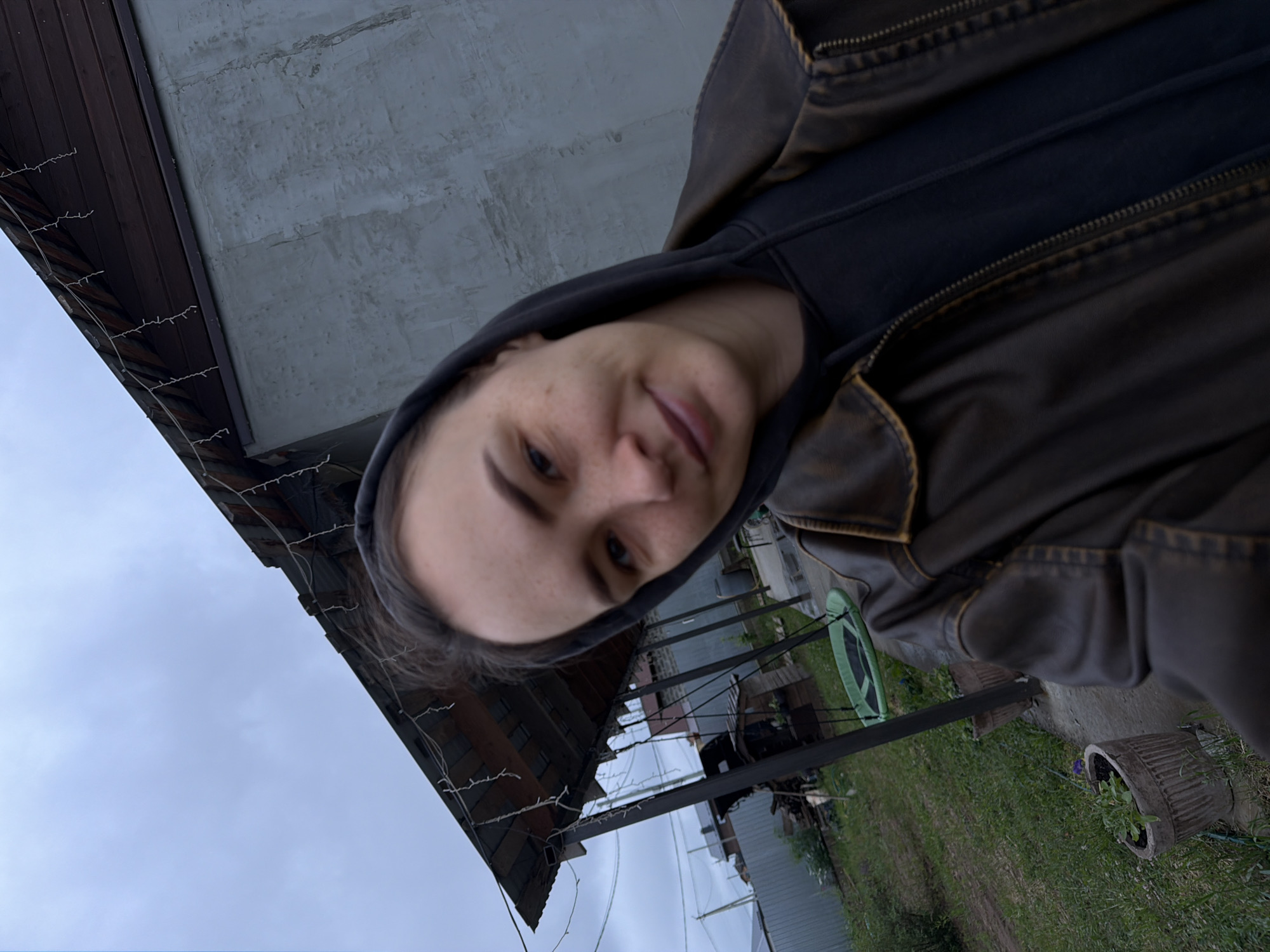 A selfie-style photograph appears to have been taken outside. The person in the foreground is wearing a dark hooded sweatshirt and a brown leather jacket. The background shows a building with a wooden roof and a concrete wall. There's an expanse of cloudy sky visible in the corner. A yard with green grass, assorted objects, and plants in pots are also visible. Based on the available information, there is insufficient information to determine whether the location is Chaikovsky, Russia.