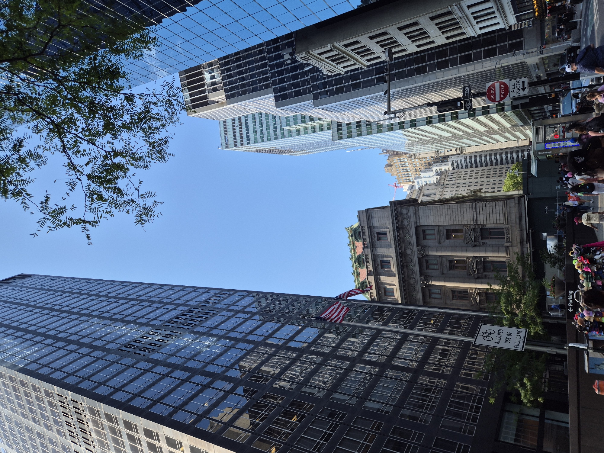 In New York City, USA, a low-angle perspective captures a vibrant street scene dominated by towering skyscrapers and a clear blue sky. In the foreground, a dark, glass-fronted building reflects the surrounding architecture and sky. To its left, a flagpole displays the American flag. To the right, a lighter, ornate building with a green-tiled roof stands out.
Further down the street, modern glass skyscrapers rise, creating a canyon-like effect. A street sign indicates "ALL TRAFFIC" with an arrow pointing right and a "DO NOT ENTER" sign is visible on a lamppost. Pedestrians are walking along the sidewalk, some browsing merchandise displayed on racks, including colorful hats. A vehicle is visible in the street. The overall impression is of a bustling urban environment on a bright day.