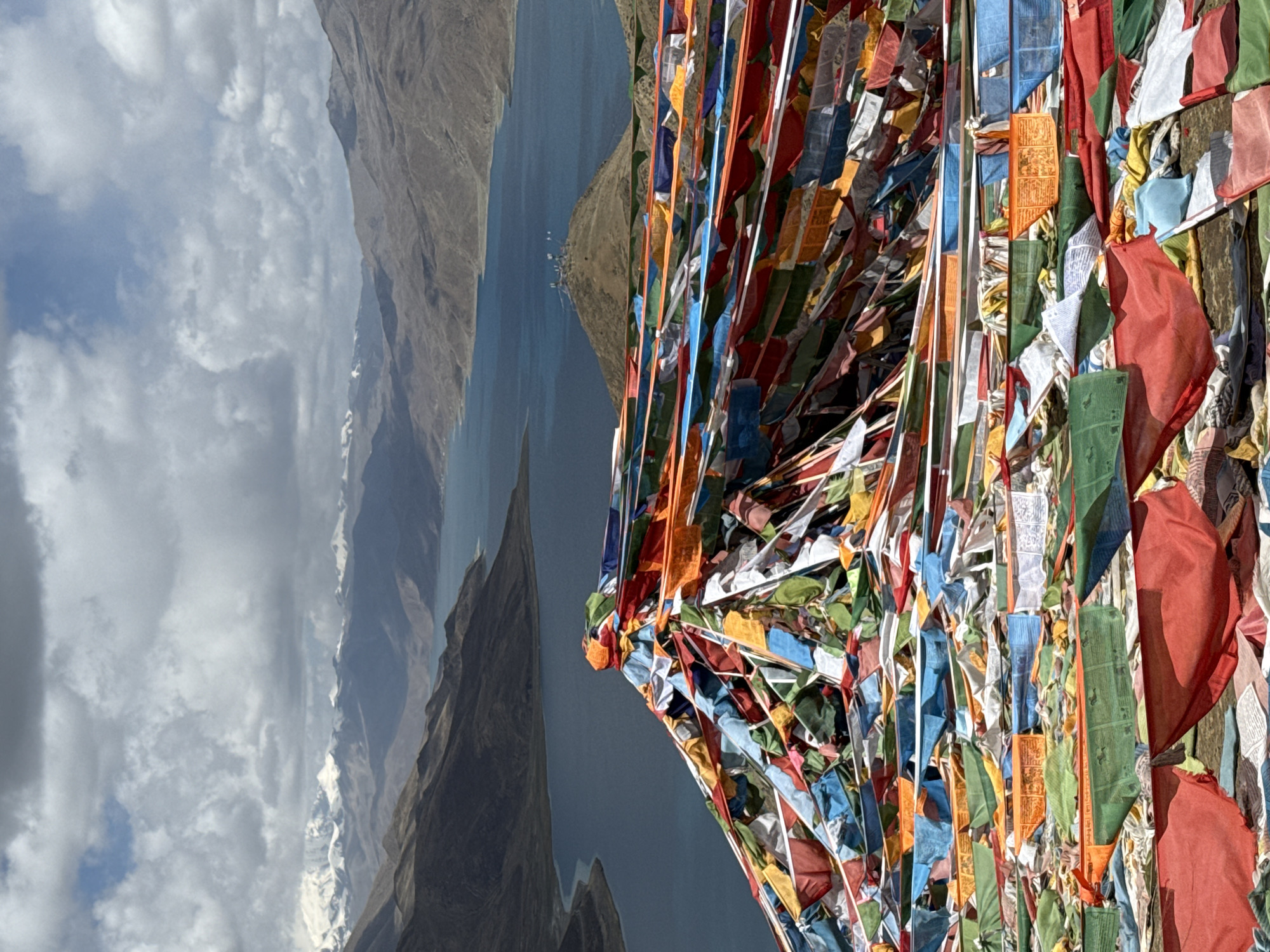 The image depicts a mountainous landscape in Qangtang town, China, with a body of water in the background. Prayer flags in various colors are in the foreground. The flags are heavily clustered together. Mountains dominate the left side of the image, while a clouded sky appears in the distance.