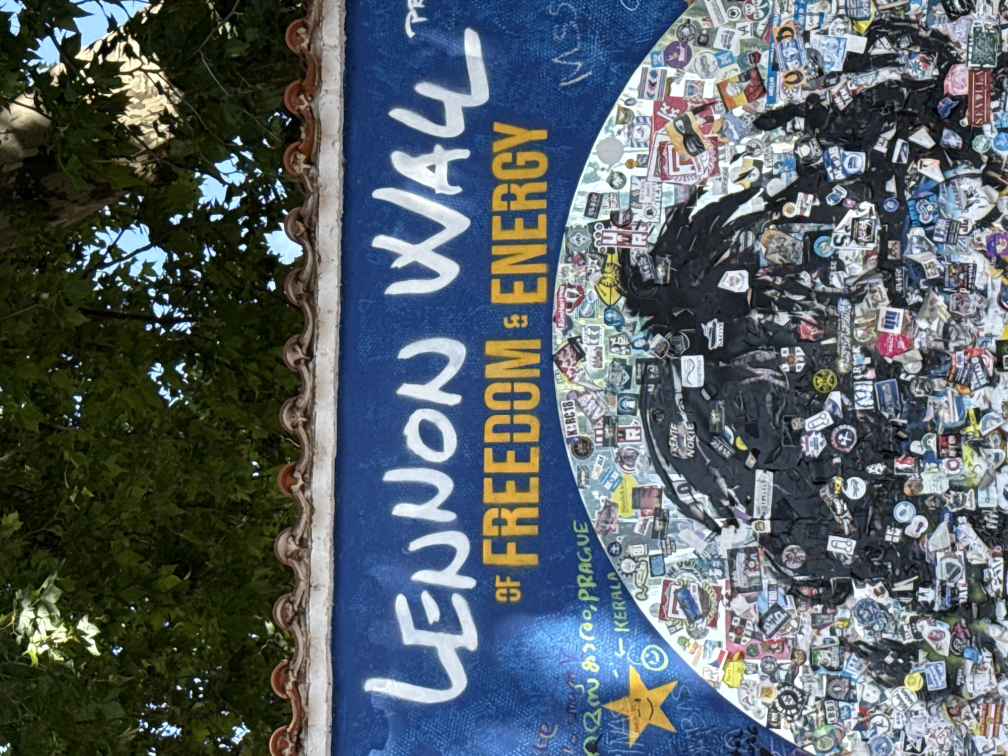 The image shows a section of the Lennon Wall in Prague, Czechia. The wall is covered in graffiti, paintings, and numerous stickers. The top of the wall features white lettering that reads "LENNON WALL" and below it, in yellow, "OF FREEDOM & ENERGY". Below this, the wall is a mosaic of colorful stickers and smaller drawings, with a prominent black silhouette of what appears to be a bird or a winged creature in the center. The lower portion of the image shows more stickers and some Malayalam script, along with the word "PRAGUE" and "KERALA". In the upper part of the image, green foliage from trees hangs over the wall, and a blue sky is visible through the leaves. The wall itself is painted a vibrant blue.