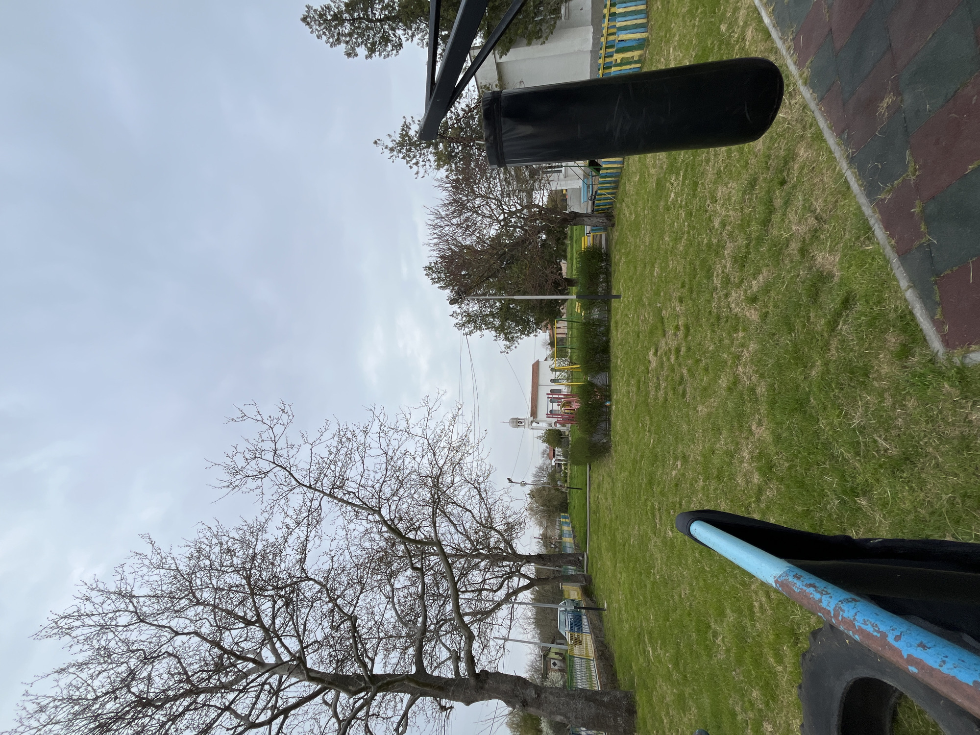 The image shows a grassy area in Polski Izvor, Bulgaria, with trees lining the periphery. A brick-patterned walkway borders the grass. Overhead, there are elements of playground equipment: a black cylindrical object hanging from a metal frame and a blue-painted metal bar with a black object. In the background, a white building with a red roof is visible. The sky is overcast.