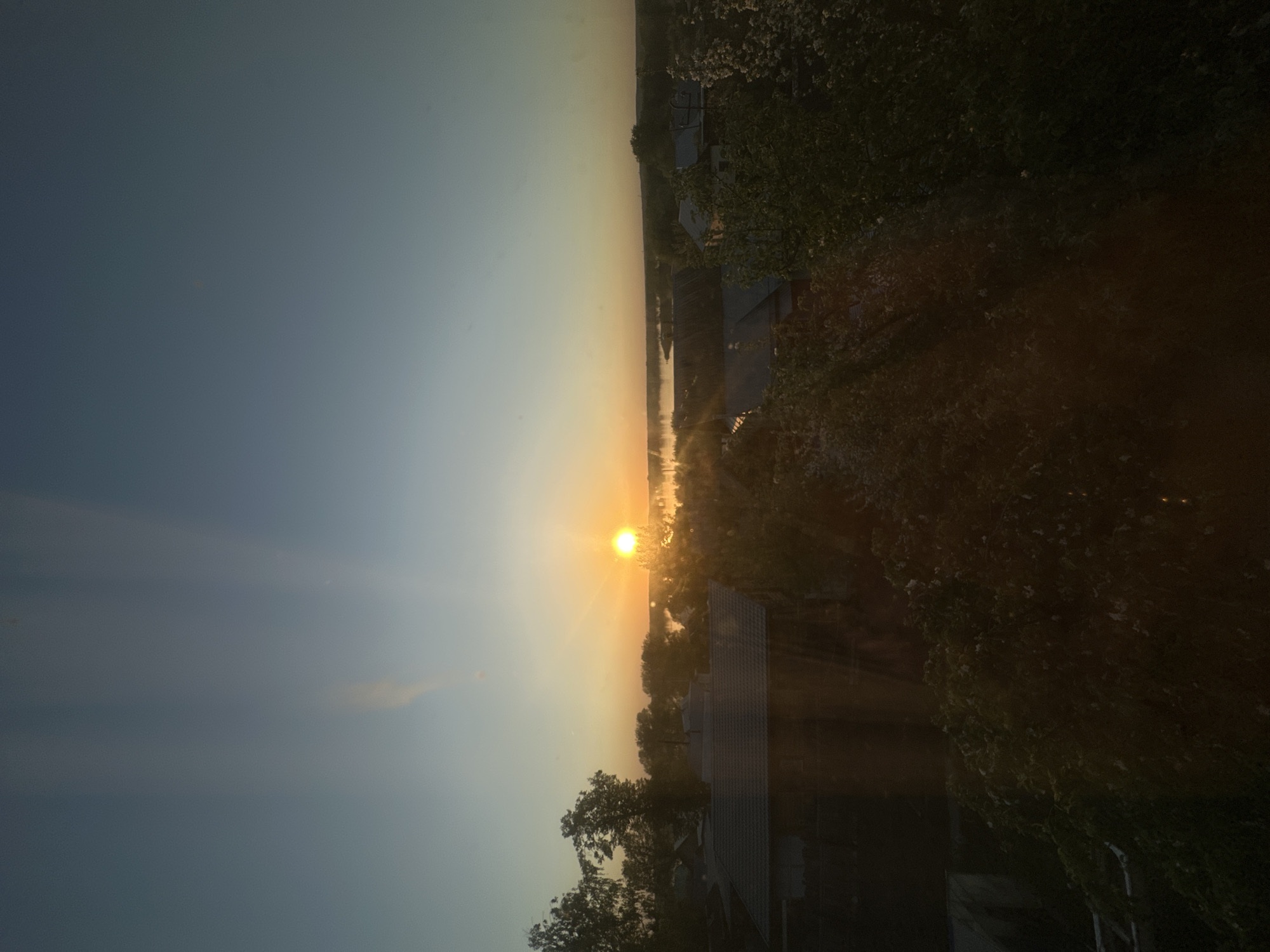 The image shows a sunrise over Chaikovsky, Russia. The sun is centrally located in the horizon, with bright light rays emanating from it. To the left of the sun is a blue sky, and to the right are buildings and vegetation. A river or other body of water is visible in the distance. Some trees appear to be in the foreground on the lower part of the frame. The perspective is from an elevated position.