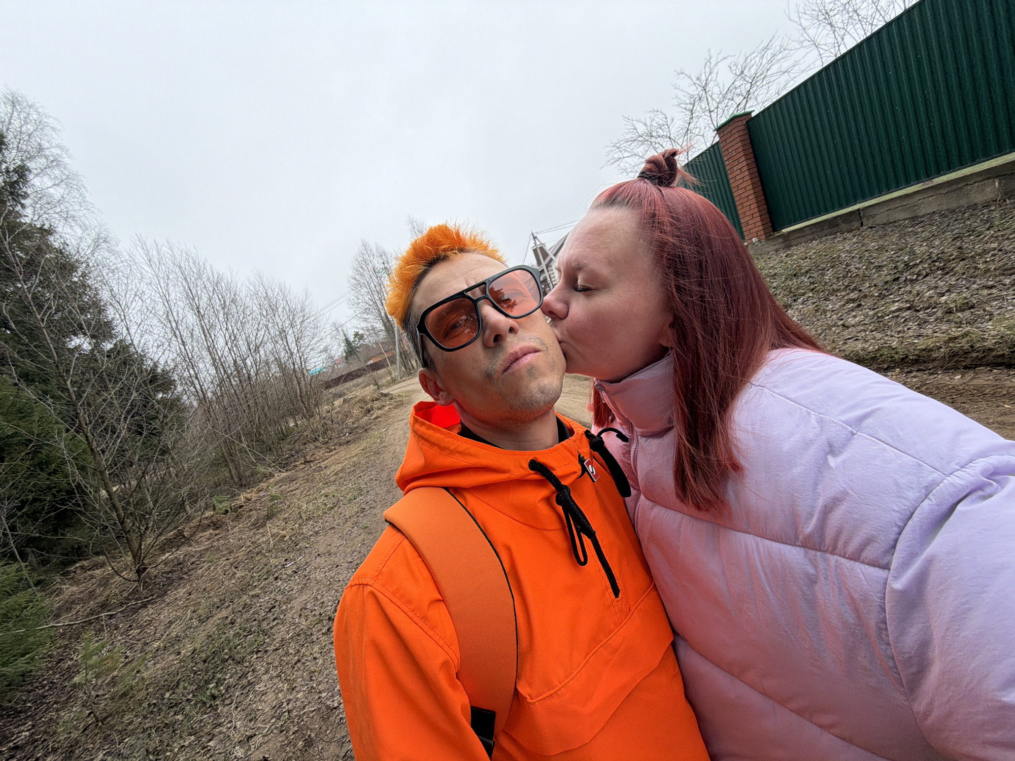 This outdoor, wide-angle photograph from Chaikovsky, Russia, features a couple in what appears to be late winter or early spring. The sky is uniformly overcast and bright grey, indicating a cloudy daytime.
In the foreground, a man is prominently positioned on the left, looking directly at the camera. He has bright, spiky orange hair and wears large black sunglasses with orange-tinted lenses. He is dressed in a vibrant orange hooded anorak with black drawstrings and has an orange backpack strap visible over his right shoulder. His expression is neutral or slightly serious.
Leaning in from the right to kiss his left cheek is a woman. Her eyes are closed, conveying affection. She has long, reddish-brown hair styled in a bun on top of her head and is wearing a light lavender or pale pink puffy jacket.
The background features an upward-sloping landscape covered with dry leaves and bare deciduous trees, reinforcing the impression of a cold season. To the far right, a dark green corrugated metal fence runs along the top of the slope, accompanied by a brick pillar. No discernible text is visible anywhere in the image. The setting suggests a natural or semi-rural environment.