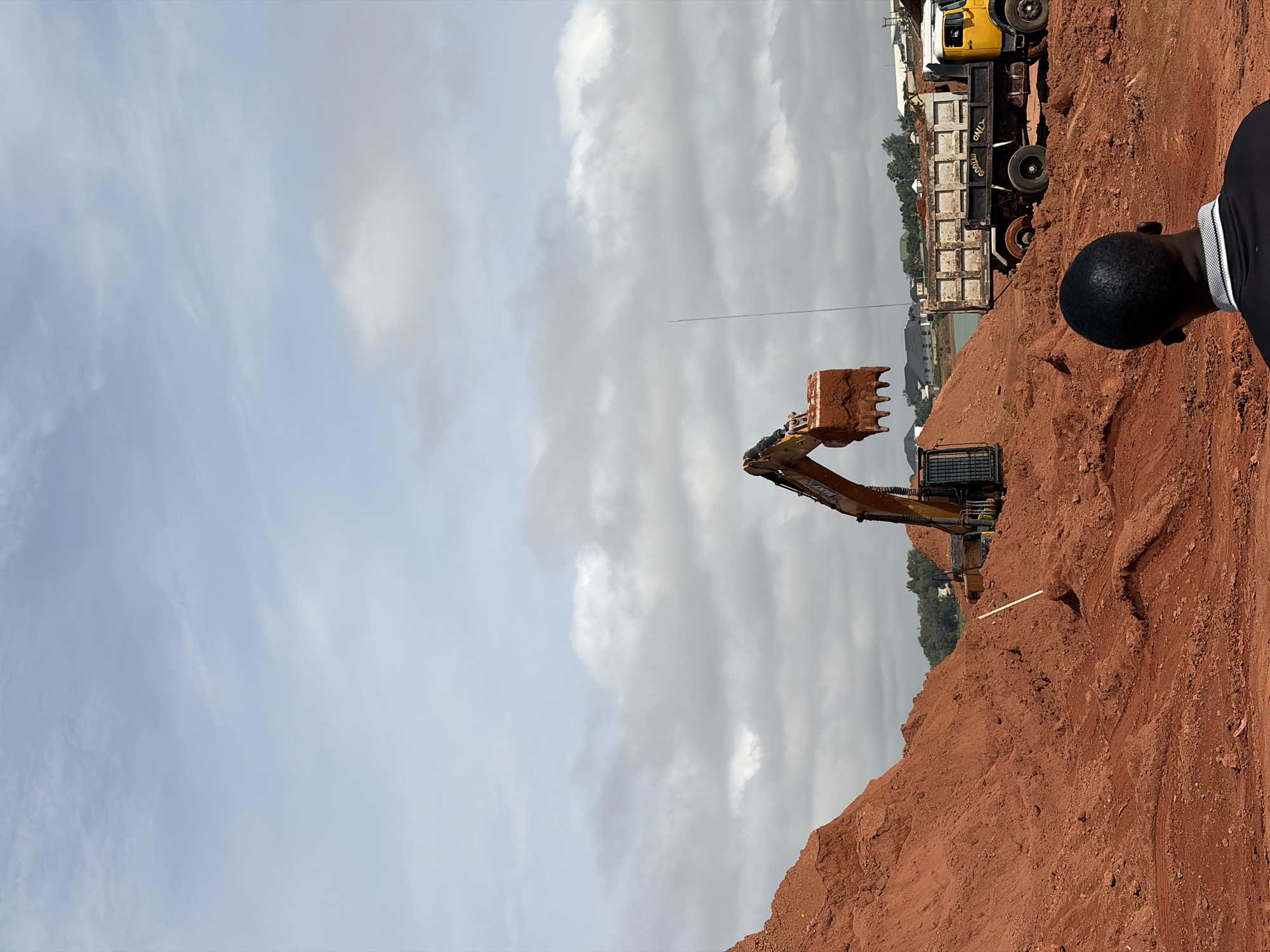The image depicts a construction site in Kwang, Nigeria, dominated by large mounds of red soil. An excavator is actively digging into one of the mounds, with its bucket full of dirt. A yellow dump truck with the words "GOODS ONLY" written on its side is parked nearby, presumably ready to be loaded. In the foreground, a man with short, dark hair is seen from behind, looking towards the excavation. The background features some buildings and trees, indicating the presence of a settled area near the construction zone. The sky is partly cloudy, with a light blue hue.