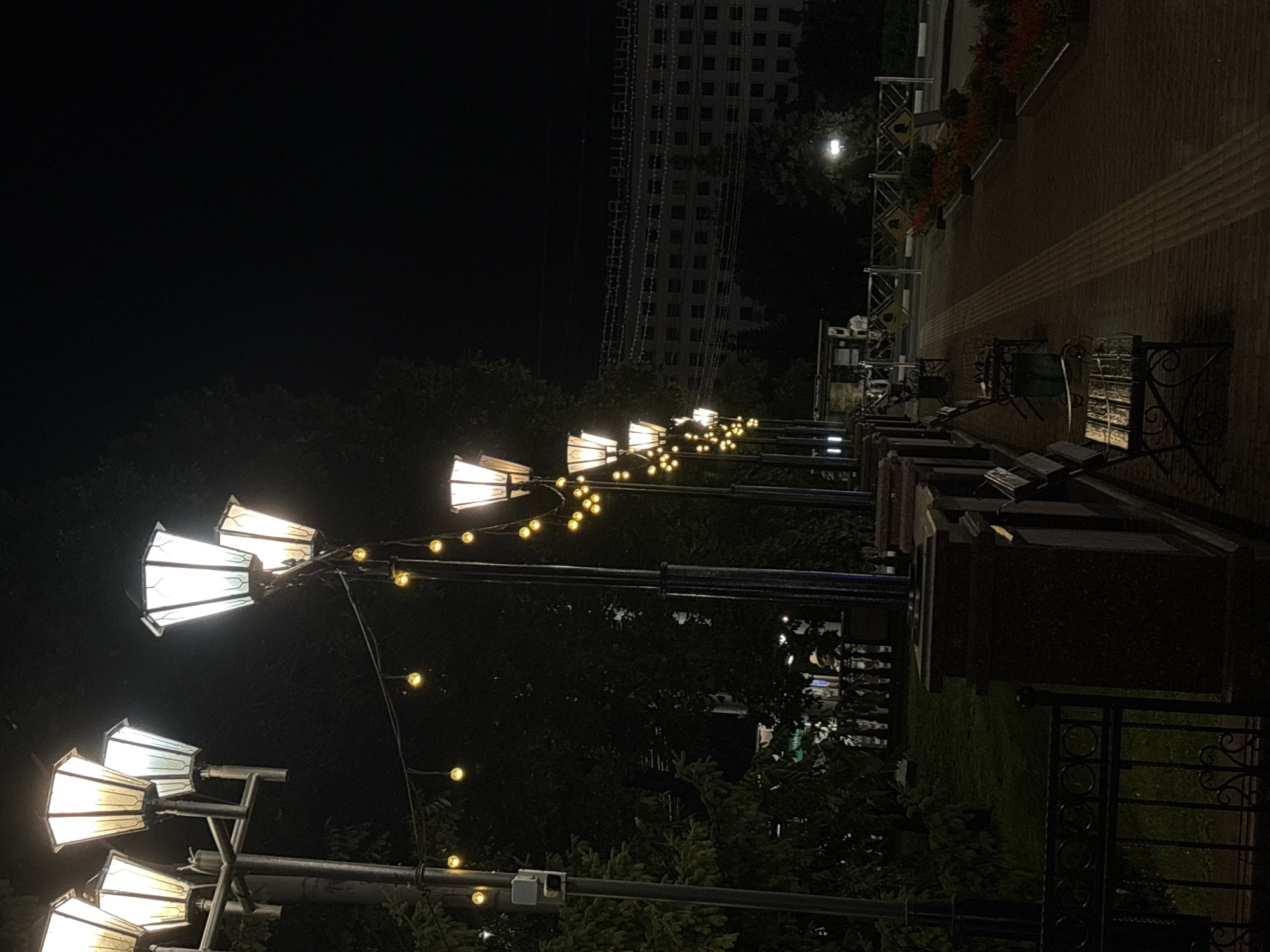 The image depicts a nighttime scene in Vitebsk, Belarus, likely along an illuminated pathway. In the foreground, a row of ornate lampposts with white, hexagonal shades line the left side of the frame, casting a warm glow. String lights with small, yellow bulbs are also visible, draped between some of the lampposts. A dark fence separates a grassy area with trees from the pathway.
The pathway itself is paved with bricks and features a tactile warning strip. Several park benches are placed along the pathway, some occupied by indistinct figures. In the background, a large, multi-story building with many windows is illuminated and appears to have signage with Cyrillic lettering. Red flowers are planted in beds along the right side of the pathway, adding a touch of color to the dark scene. The overall atmosphere is serene and well-lit, suggesting a public space open for evening use.