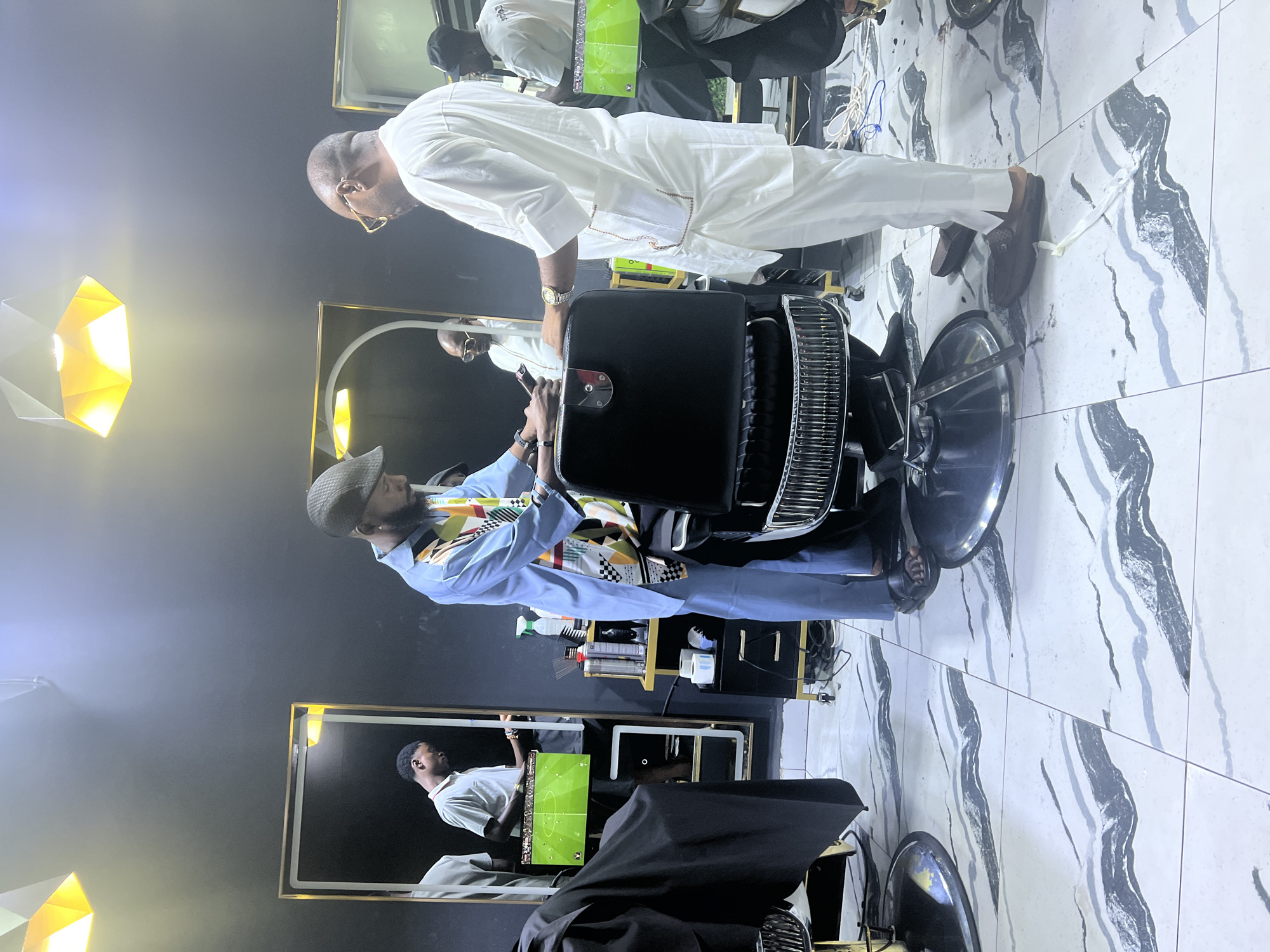 The image shows the interior of a barbershop in Lugbe, Nigeria. A man in a light blue shirt and hat is seated in a barber's chair. Another man, dressed in white, stands behind the chair, seemingly adjusting or preparing the chair for the customer. A barber is also standing in the reflection of the mirror. A television screen is visible in the background, and barbering supplies can be seen on a shelf. The floor is tiled with a grey-and-white marble pattern. A soft box is visibile in the corner of the room providing lighting.