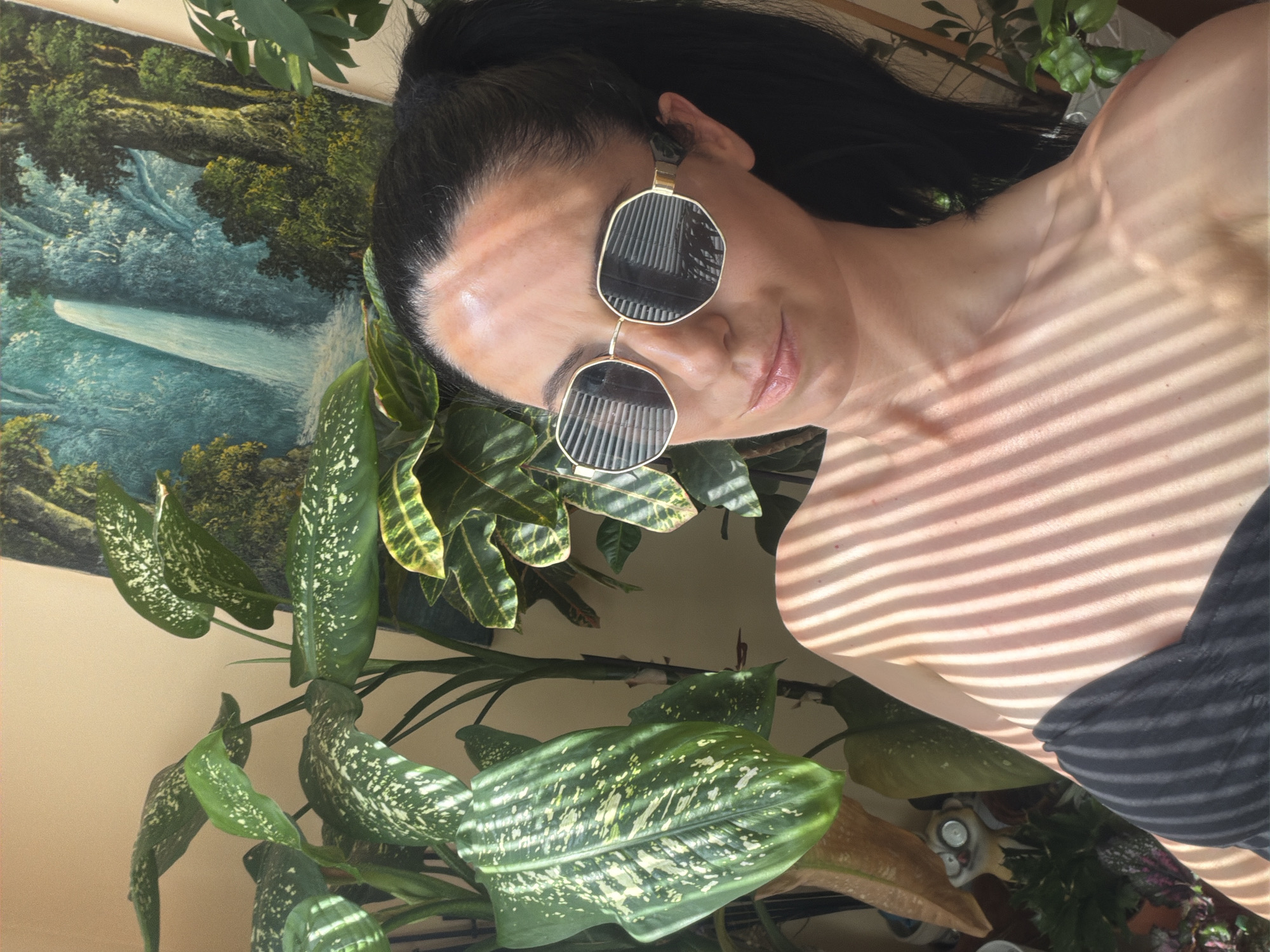 A woman with black hair pulled into a ponytail, wearing large, octagonal sunglasses with gold frames, poses for a selfie. She is surrounded by lush green plants, and a painting of a waterfall is visible in the background. Sunlight casts striped shadows across her face and chest. She is wearing a black strapless top. The scene appears to be indoors, possibly in a residential setting. There is no discernible connection to the city of Pleven, Bulgaria, nor any information about its specific location within that city.