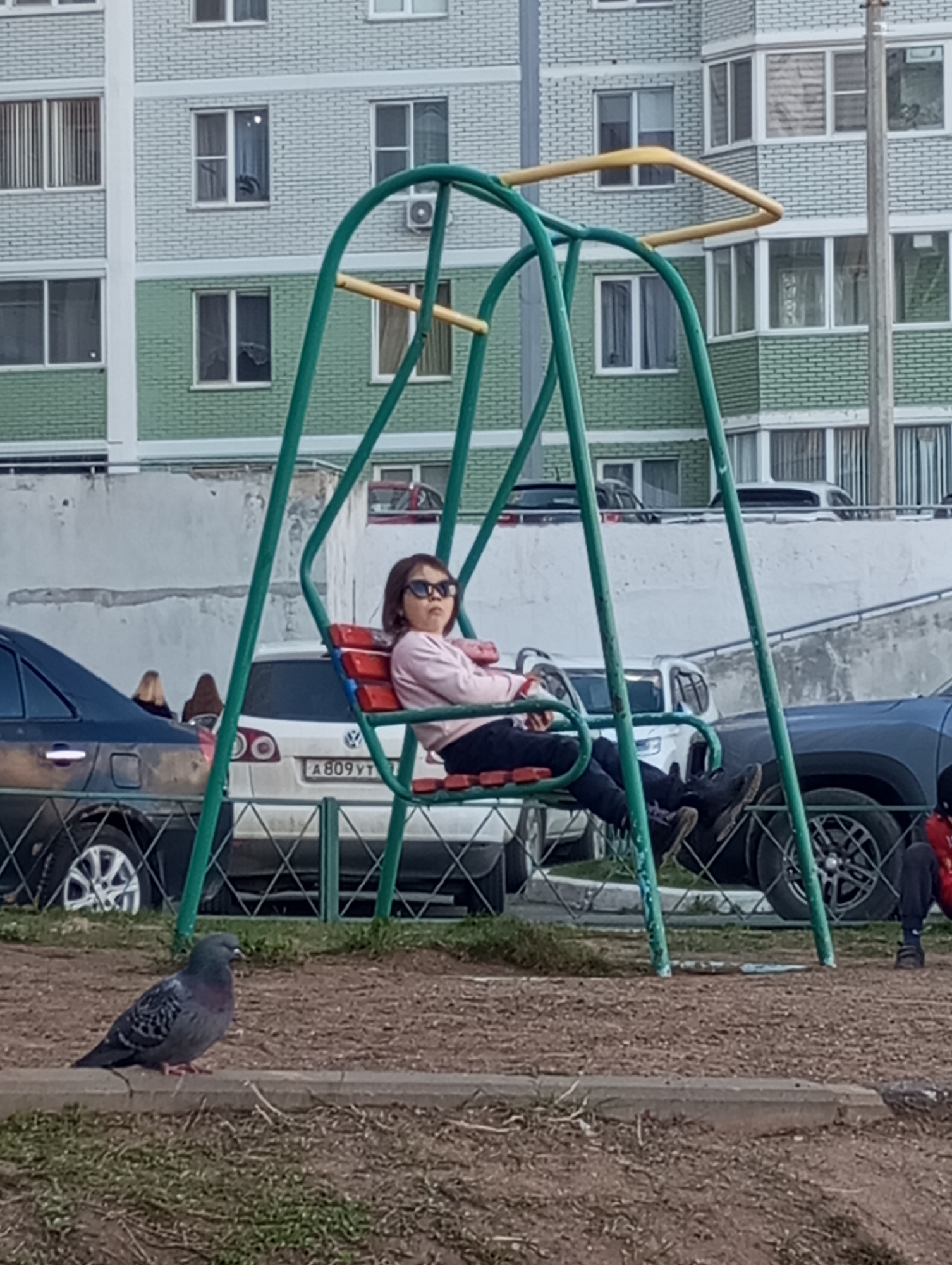 A young girl sits in a green and yellow playground swing in an urban environment. She wears sunglasses, a light pink sweater, and dark pants. The background contains a gray and green apartment building and several parked cars. A pigeon is standing on the ground in front of the swing set. The license plate visible on one of the cars reads "A809YT." Given the Cyrillic script, the location is determined to be Russia, with the registration matching that of the city of Izhevsk.