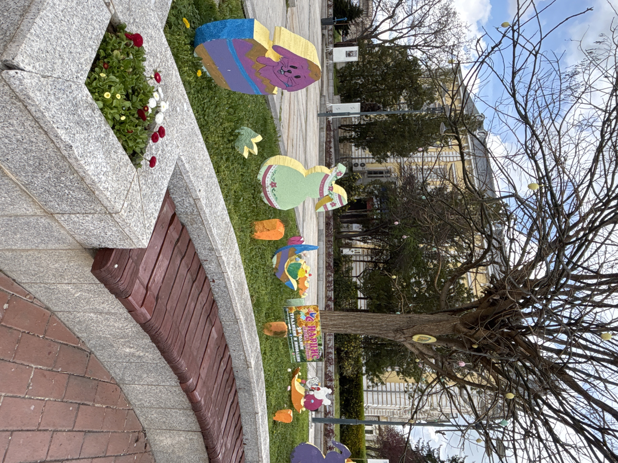 The image depicts a city park in Silistra, Bulgaria. On the left side, a raised flower bed and a bench are visible. Along the green grass area, numerous colorful, stylized, cartoonish Easter decorations are placed, including rabbits and Easter eggs. A bare tree stands on the right, with Easter eggs hung on its branches. In the background, a building with yellow paint can be seen. The sky is visible above the building and through the tree branches, indicating a daytime scene.
