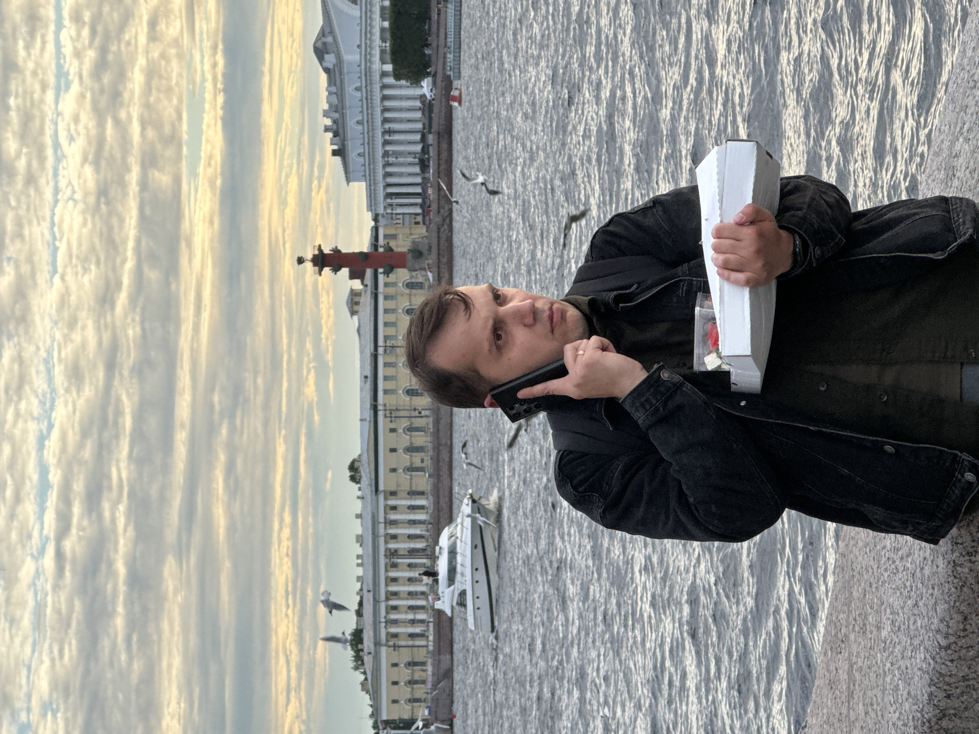A man stands on a stone embankment by a body of water, holding a white pizza box and talking on a black smartphone. He is wearing a dark denim jacket over a dark shirt and has a backpack on. The water has moderate ripples, and there are several seagulls flying in the background. Across the water, a white motorboat is visible, and further beyond that are buildings with classical architecture, including a tall red column. The sky is overcast with layers of clouds, some of which are illuminated by sunlight. The scene appears to be the Neva River in Saint Petersburg, Russia, given the iconic architecture and the spire of the Peter and Paul Fortress in the background.