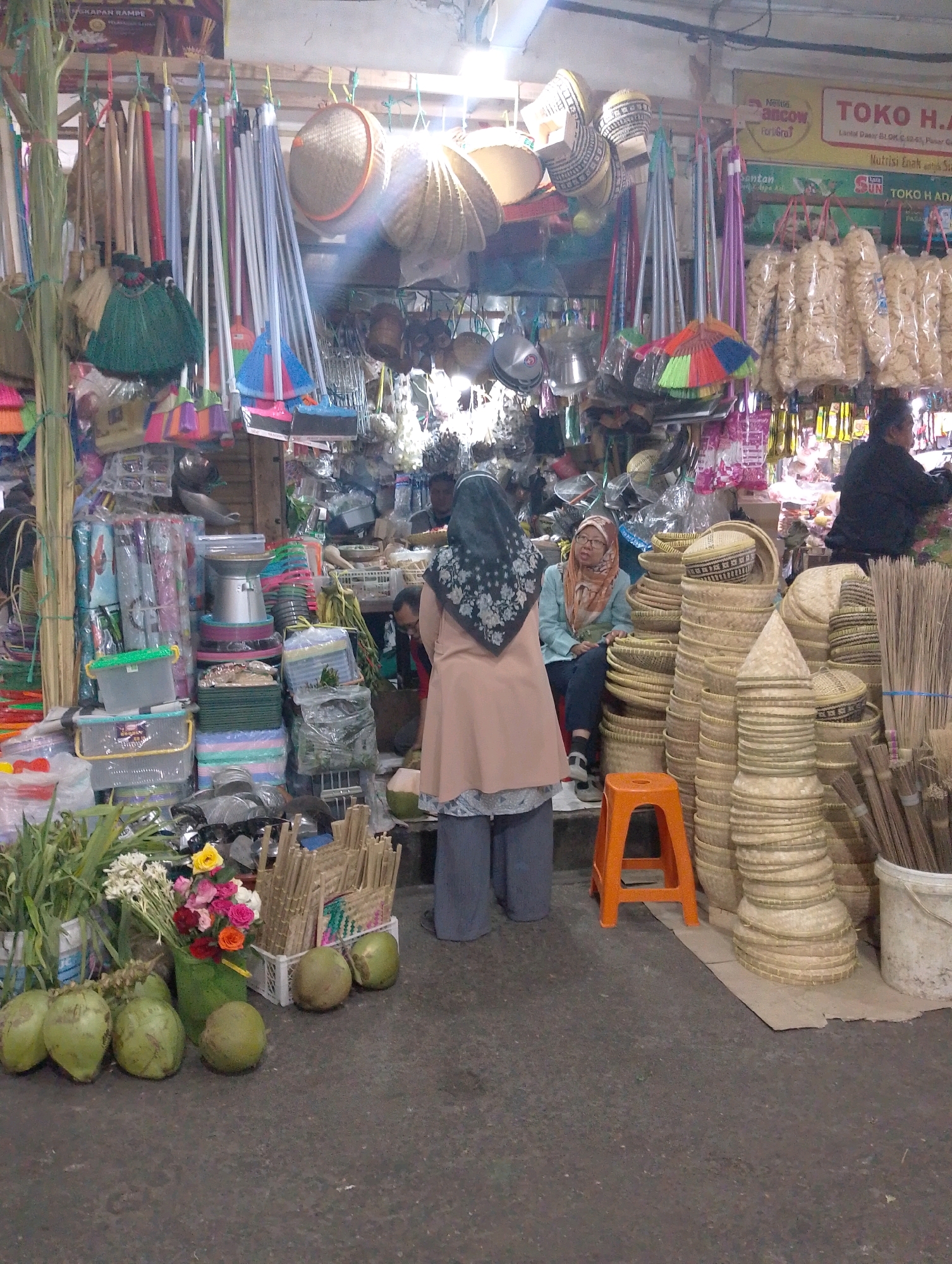 The image depicts an outdoor market stall in Lembang, Indonesia, filled with a variety of goods. The stall is packed with items ranging from household supplies to food products. In the foreground, several green coconuts are displayed on the ground, along with a vase of colorful flowers. Behind the coconuts, a stack of woven bamboo items, possibly baskets or mats, dominates the central part of the scene.
Several people are visible within the market. A person wearing a peach-colored hijab and a light blue top is seated among the bamboo goods. Another person in a dark jacket is seen behind the counter, potentially attending to customers. Two other individuals, dressed in casual clothing, are also present in the background, browsing or working.
The stall is organized with multiple levels of shelving and hanging displays showcasing a diverse inventory. Items include brooms and cleaning tools, plastic containers, packaged goods, and various woven crafts. The overall atmosphere suggests a bustling local market environment in Lembang. The ground is paved with asphalt, and the stall is covered by an overhead structure, hinting at a semi-permanent setup.