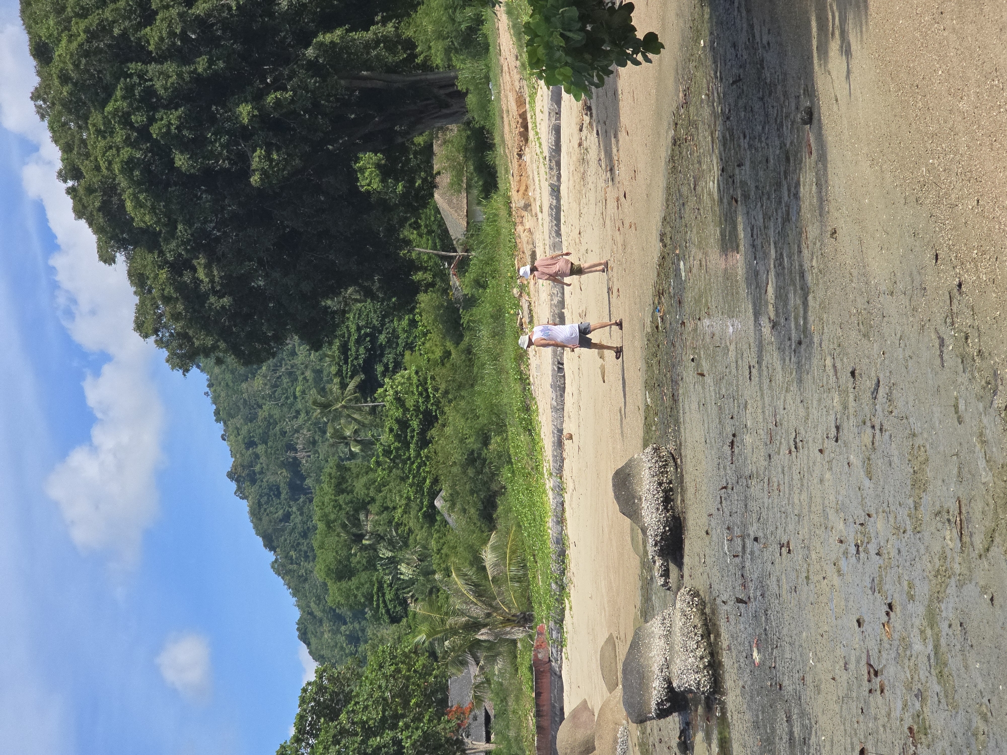 The image depicts a tropical beach scene in Baan Chaweng Noi, Thailand, under a bright blue sky with scattered white clouds. The foreground is a wet, sandy area with scattered debris and small tide pools. Several large rocks, encrusted with barnacles, are visible in the mid-ground, some partially submerged in water. Two individuals, a man and a woman, are walking away from the camera on the dry sand of the beach. The man, wearing a white t-shirt and a light-colored hat, is on the left. The woman, wearing a light-colored t-shirt and a pink hat, is slightly ahead and to the right of the man.
Behind the beach, lush green vegetation covers a sloping hillside. Palm trees and dense foliage dominate the landscape, with a few thatched-roof structures partially visible amidst the greenery, suggesting a residential or resort area. The overall environment conveys a serene, natural setting typical of a Thai island.
