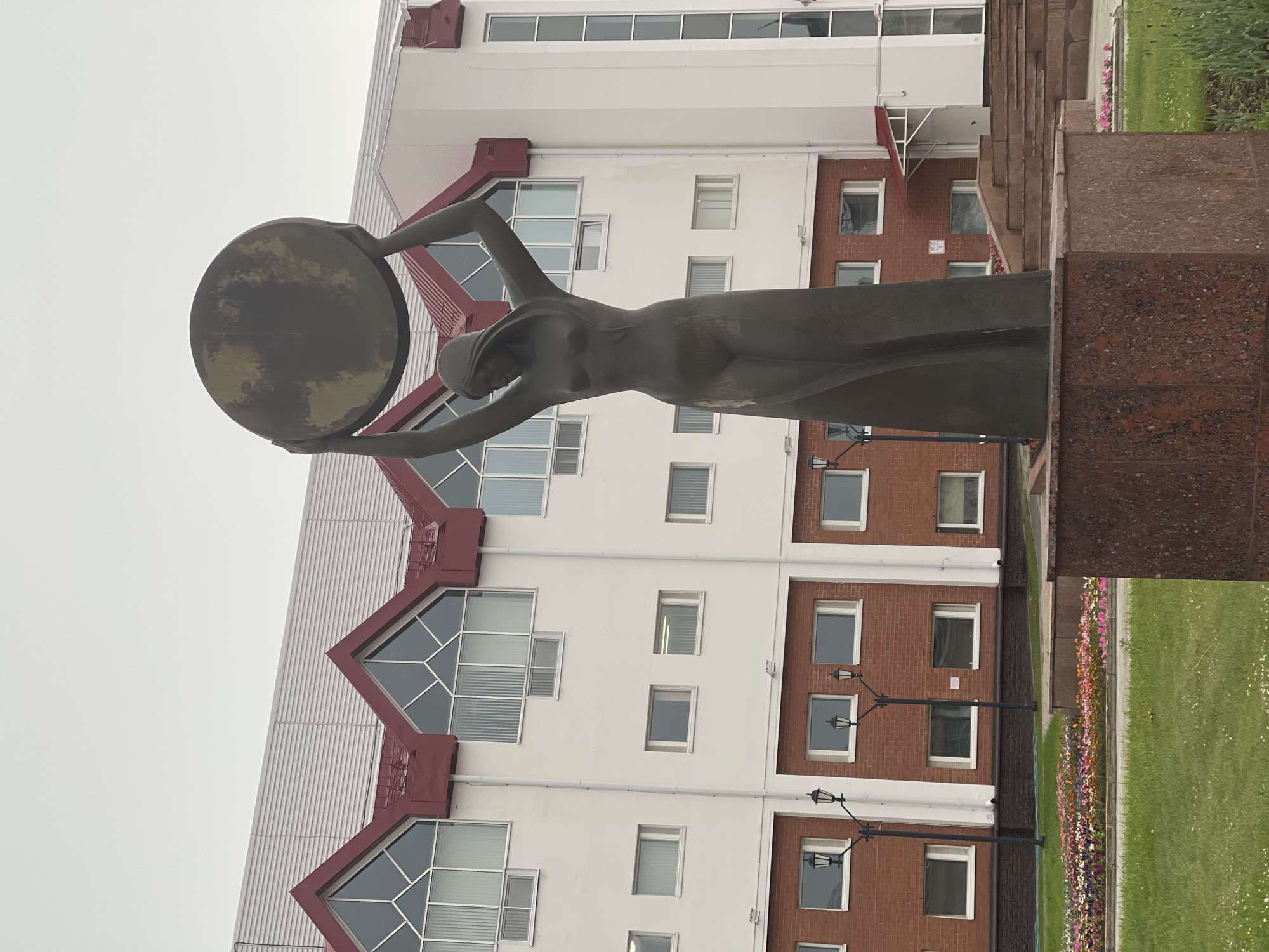 This is a vertical, eye-level, outdoor shot of a bronze statue of a woman holding a large, round disc above her head. The statue is in the foreground, to the left, and is made of dark, weathered bronze. The woman is depicted in a flowing gown, with her arms outstretched, holding the disc, which has a mottled, reflective surface.
Behind the statue is a modern building with white and red-brown brick facades. The building has many windows, some large and angular with geometric divisions, and others smaller and rectangular. The overall architectural style appears to be functional and contemporary. In front of the building and to the right of the statue, there is a neatly kept lawn with a colorful flowerbed running along its edge. Several lampposts with traditional designs are also visible in front of the brick section of the building.
The sky is overcast and a uniform light gray, suggesting a cloudy or possibly rainy day. The lighting is diffused, casting soft shadows. The scene appears to be a public space, possibly a park or plaza in front of an institutional building in Khanty-Mansiysk, Russia, given the location context. There are no people visible in the frame, and the statue's pose and the surrounding environment evoke a sense of solemnity or perhaps a tribute to an abstract concept. No text is visible in the image.