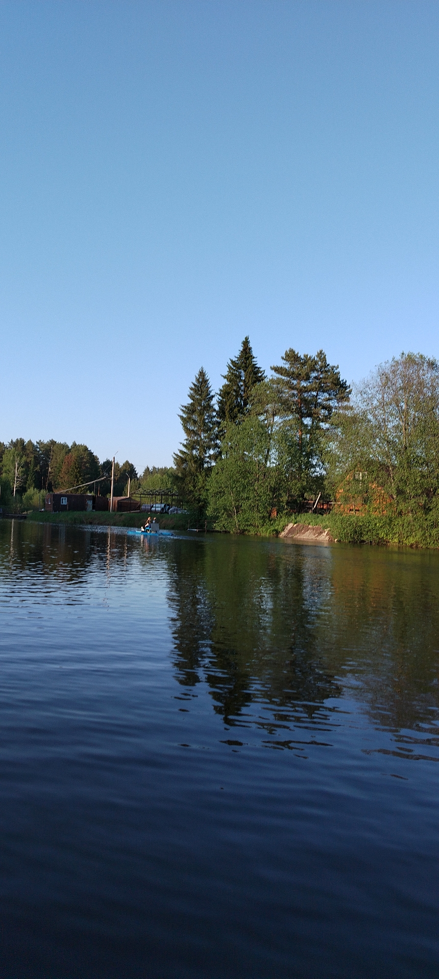 The image shows a lake with reflections of trees and sky in Prikamskii, Russia. A small blue paddle boat with two figures is visible in the middle ground. A building, trees, and a structure, possibly a bridge, are on the shoreline. The sky is clear and blue. The water is dark with gentle ripples.