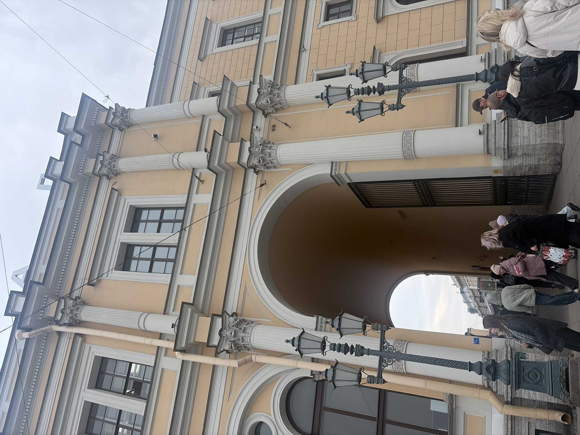 The image displays the eastern archway of the General Staff Building, located on Palace Square in Saint Petersburg, Russia. The multi-story classical structure features a light yellow facade with white window trim and cornices, and prominent fluted white Corinthian columns with ornate capitals. A large, deep yellow-orange arched passage dominates the lower center, with a dark metal gate partially visible inside. Dark-framed rectangular and arched windows are present across the building's face.
In the foreground, two dark grey, ornate lampposts, each equipped with three lanterns, stand on the grey paved sidewalk. Approximately ten to fifteen individuals are visible on the sidewalk and within the archway. Most are dressed in various winter coats and some wear hats. Actions include walking, standing, and observing. One person on the left foreground looks upwards. Another individual in the mid-ground, wearing a black jacket and backpack, is observed looking at a mobile phone. Overhead electrical wires are strung across the scene against an overcast, light grey sky. A beige downspout runs vertically along the left side of the building. A white, angular 'A' shaped architectural element or sign is visible on the building's rooftop, silhouetted against the sky.