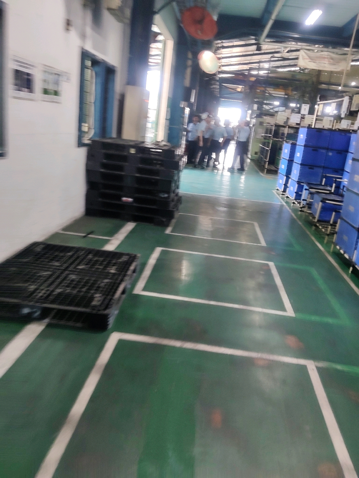 The image depicts an indoor industrial setting, likely a warehouse or factory floor, with a green-painted concrete floor marked with white lines. In the foreground, a black plastic pallet is visible, and further back, a stack of similar pallets is present. Several individuals, appearing to be employees wearing light-colored shirts and dark pants, are gathered in the background. To the right, numerous blue storage bins are stacked high on shelves. The environment is illuminated by overhead fluorescent lighting. The scene is located in Mekarmukti, Indonesia.