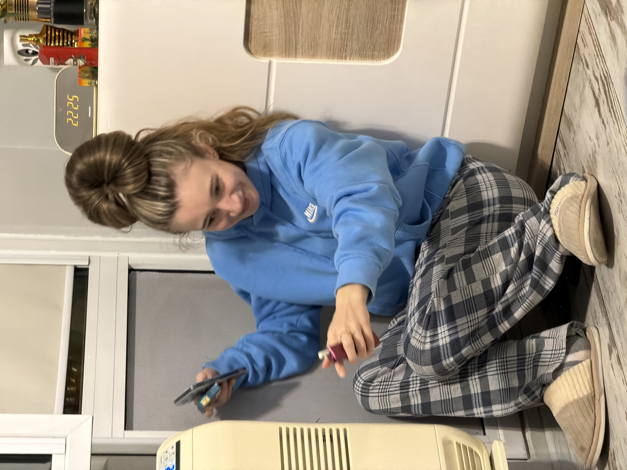 A young woman with her hair in a bun is squatting on a floor, wearing a blue Nike hoodie and plaid pajama pants with slippers. She is holding a phone in her left hand and a pink tube in her right. Behind her is a white wall with a wooden accent and a digital clock reading 22:25, along with some bottles on a shelf. To her left, there is a window with a screen and a white frame. In front of her, there is a beige-colored heater. The location cannot be determined as Plovdiv, Bulgaria.