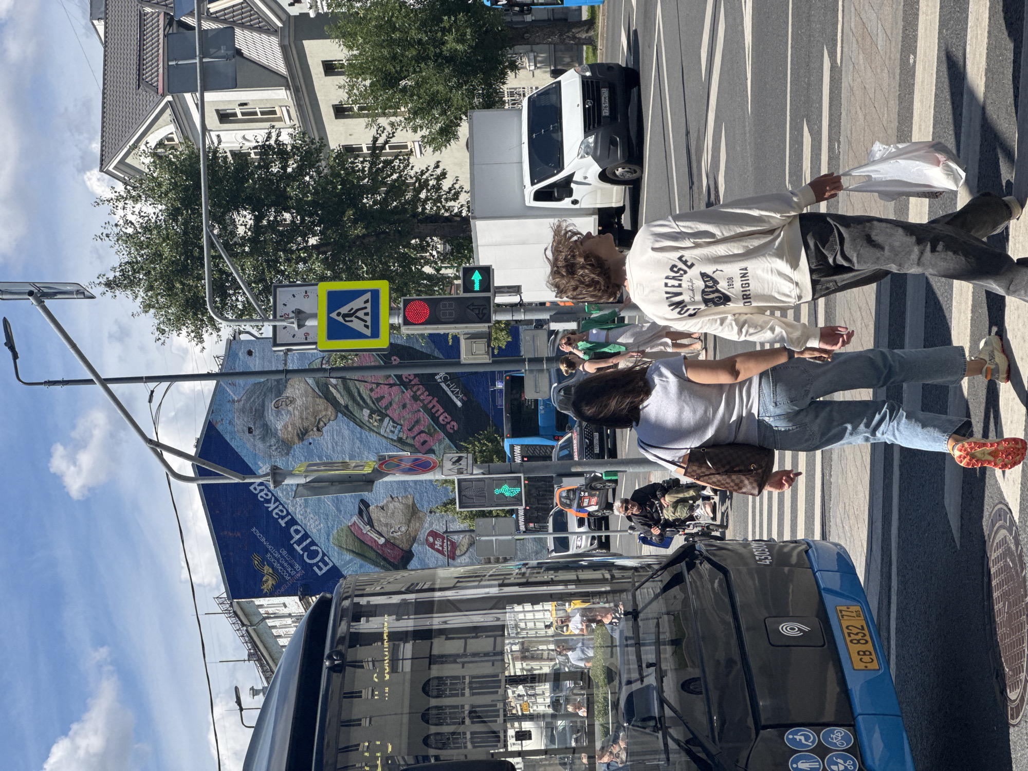 This image depicts a street scene in Moscow, Russia, likely during daytime with clear skies. The foreground features the rear of a blue bus with Cyrillic text and a license plate reading "CB 832 77 RUS". A pedestrian crossing is visible on the street.
In the middle ground, a woman in a white sweatshirt with "CONVERSE ORIGINAL 1908" printed on it and dark pants is walking, holding a white plastic bag. She is on the pedestrian crossing. To her left, another person is walking, and behind them, a traffic light shows a green pedestrian symbol. Further back, a white van is stopped. A large mural or billboard featuring faces and Cyrillic text, including "ЕСТЬ такая" and "РОССИЙСКОЕ ВОЕННО-ИСТОРИЧЕСКОЕ ОБЩЕСТВО", is mounted on a building.
In the background, a building with a sloped roof and windows is visible, along with trees. The overall impression is a bustling urban environment. The image is oriented vertically, with the sky and buildings appearing at the top, and the street and bus at the bottom.