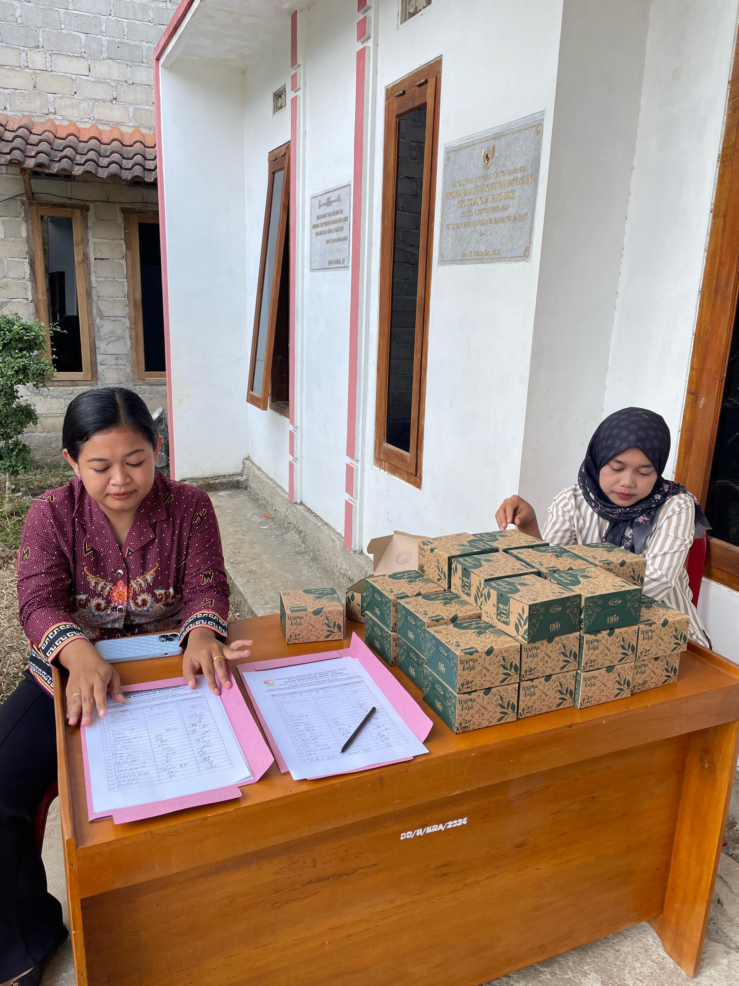 In Kagungan Ratu, Indonesia, two women are seated at a wooden desk outside a building. The woman on the left, wearing a patterned maroon shirt and black pants, is looking down at papers on the desk, with her hands positioned as if writing or reviewing. A light blue smartphone is visible in front of her. The woman on the right, wearing a striped shirt and a black hijab, is looking towards the papers on the desk. A stack of green and brown boxes with decorative designs is arranged on the desk to her right. Two pink folders containing documents are spread across the desk, with a pen resting on one of them. The building in the background is white with pink trim and features wooden doors and windows. A plaque is affixed to the building's exterior wall. The ground is paved with concrete, and dry grass is visible to the left of the desk.