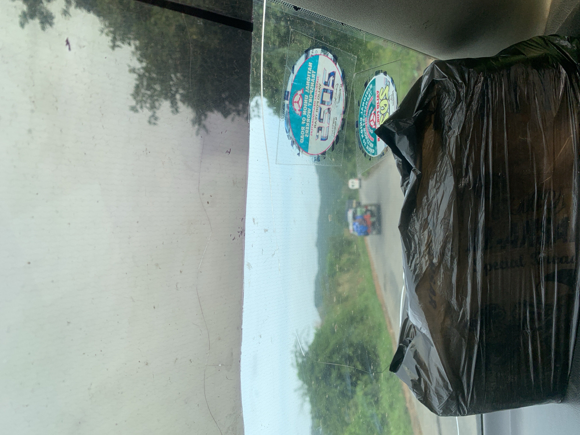 An exterior, eye-level, medium shot shows a view from inside a vehicle looking forward through the windshield. The windshield is dirty and scratched, with a black plastic bag obscuring the lower part of the view. Outside, a road curves through a lush, green landscape with trees and hills under a cloudy sky. A few vehicles are visible on the road, including one with people in the back. Two stickers are visible on the windshield: one at the top reads "NATIONAL UNION OF ROAD TRANSPORT WORKERS 2505 VALID FOR YEAR", and a second one below it has similar text and a logo. The location is Assob Hausa, Nigeria.