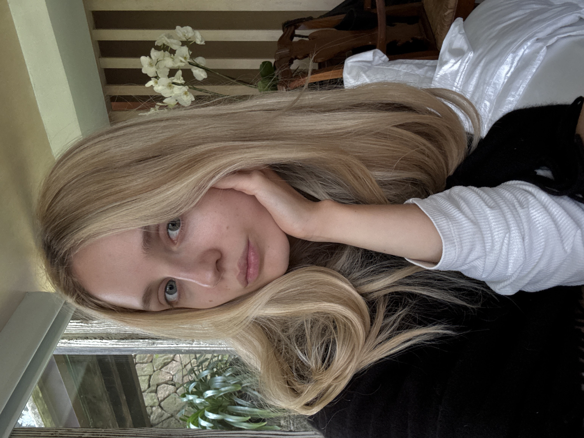 A young woman with long blonde hair and blue eyes is shown in a close-up, head-and-shoulders portrait. She is resting her chin on her hand, and her gaze is directed at the camera. She is wearing a black garment over a white ribbed shirt. In the background, a window with a view of greenery and stone structures is visible on the left. On the right, a vase with white orchids sits on a chair with decorative wooden carvings. The background also features vertical stripes of light brown and cream. The lighting suggests an indoor setting, possibly a home or a cafe, during daylight hours. There is no visual information to suggest the location is in Ciudad de México, Mexico.